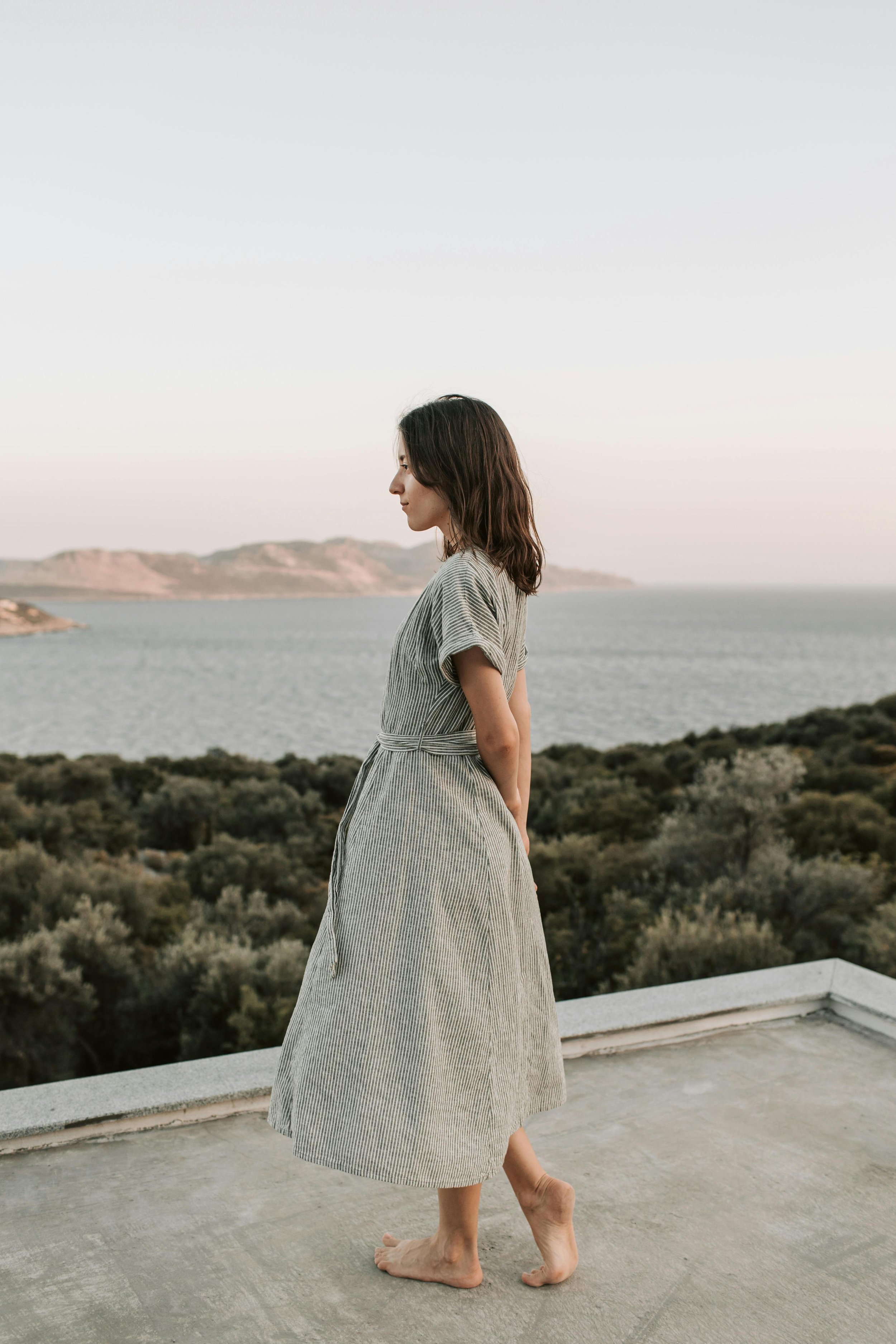 A woman with shoulder-length dark hair stands barefoot on a rooftop, looking to the left, with a scenic view of water and distant hills in the background.