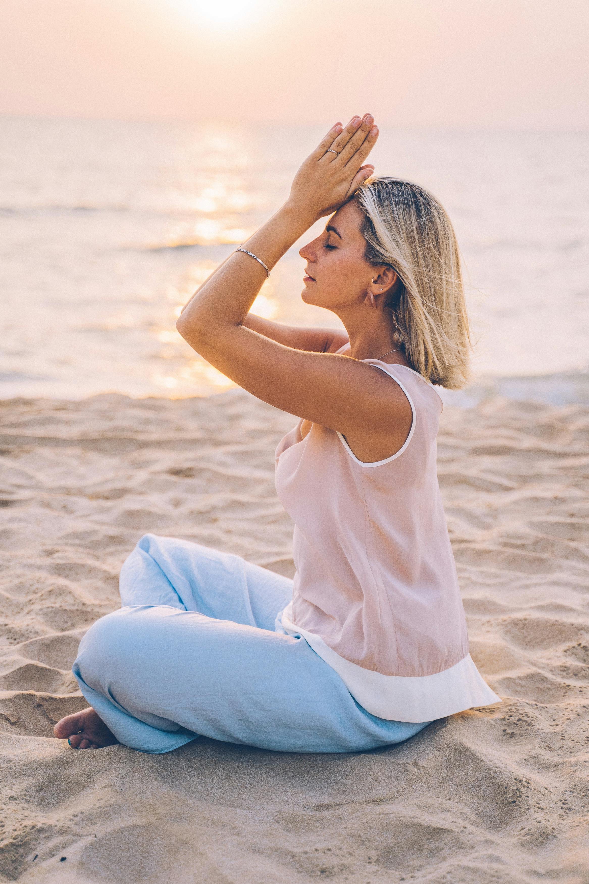 A woman sitting cross-legged on the sandy beach at sunset, meditating with her hands pressed together over her forehead.