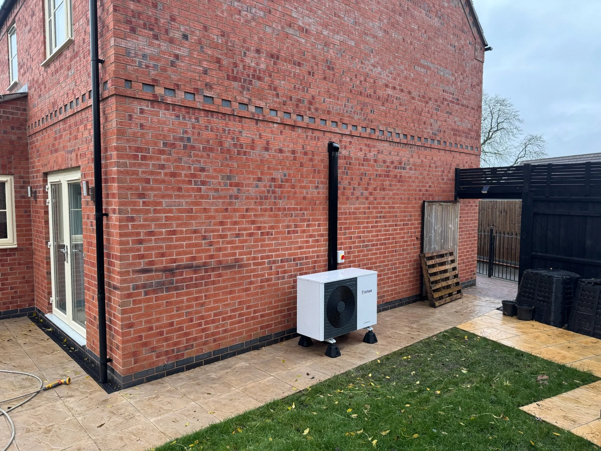 Backyard with brick house wall, black rain gutter, white Vaillant heat pump, wooden pallet, black fence, and potted plants on yellow tiled patio, adjacent to grass lawn.
