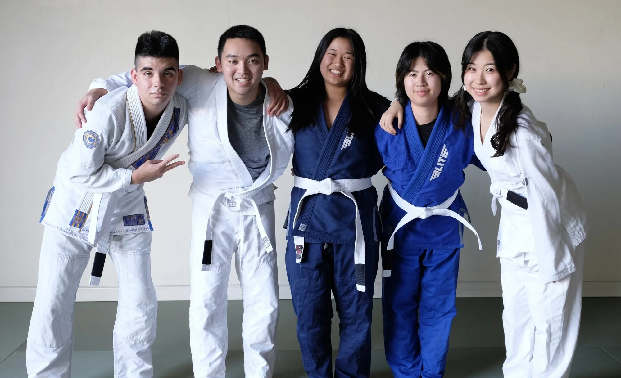 Five Budo Scholar students in gis standing together on the mat, arms around each other's shoulders