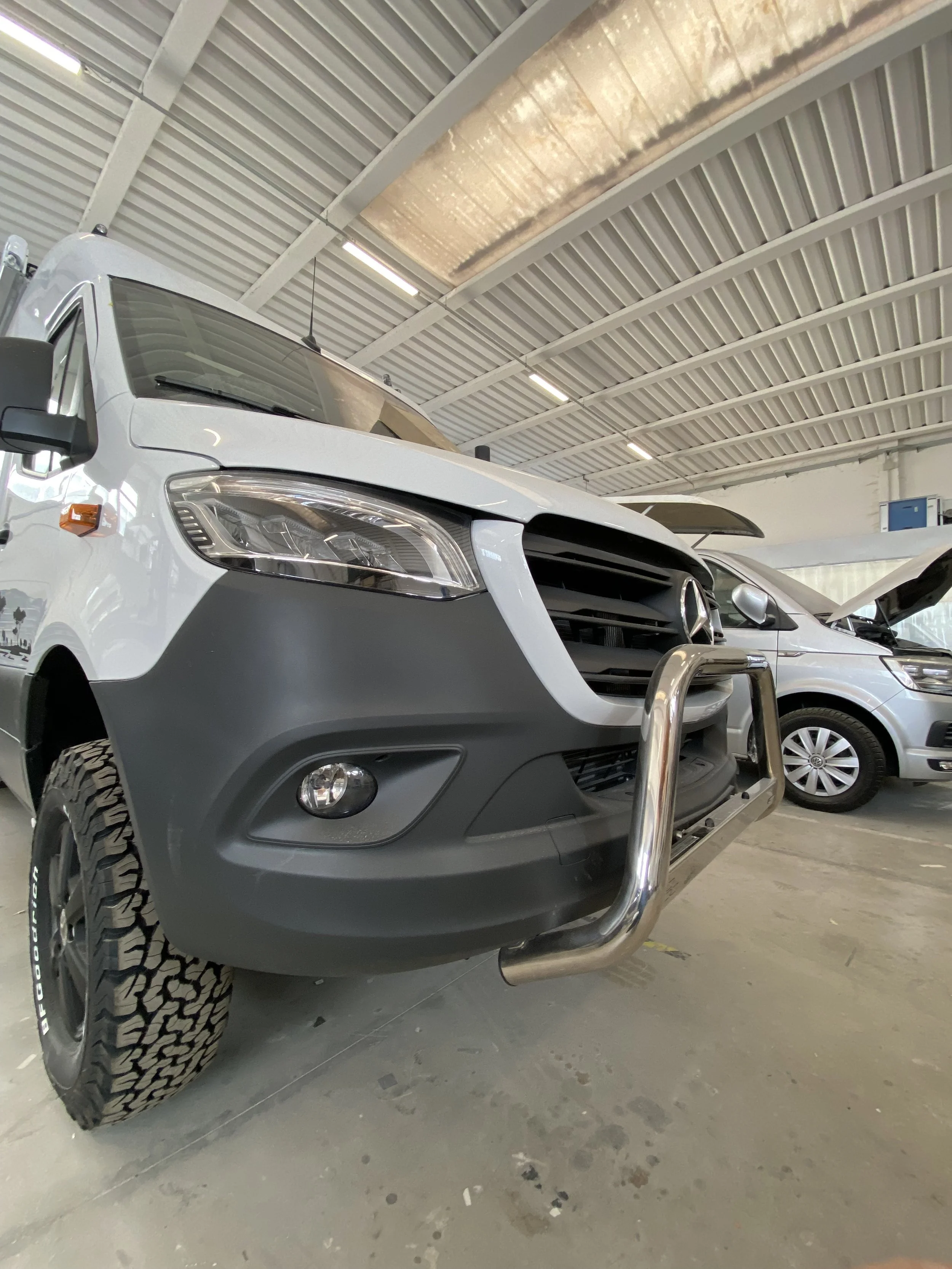 Veicolo bianco con protezione metallica anteriore e pneumatici da fuoristrada parcheggiato in un magazzino o garage con altre auto sullo sfondo.