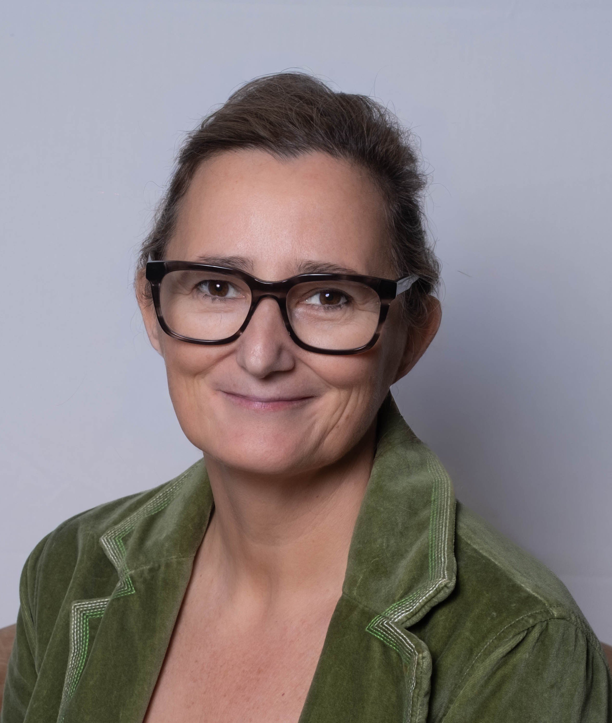 A woman with short brown hair, wearing large black glasses and a green blazer, smiling in front of a plain, light gray background.