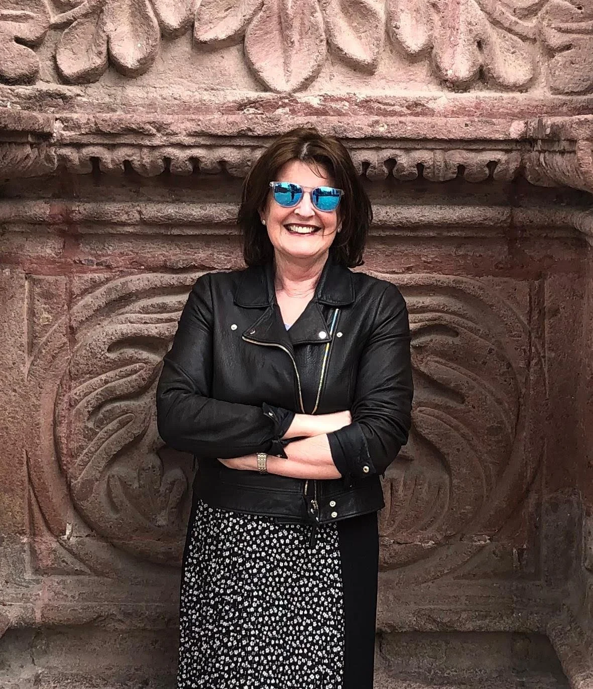 A smiling mature woman with short, wavy, brown hair wearing pearl earrings, a dark pink blazer, and a chunky black necklace against a plain light background.