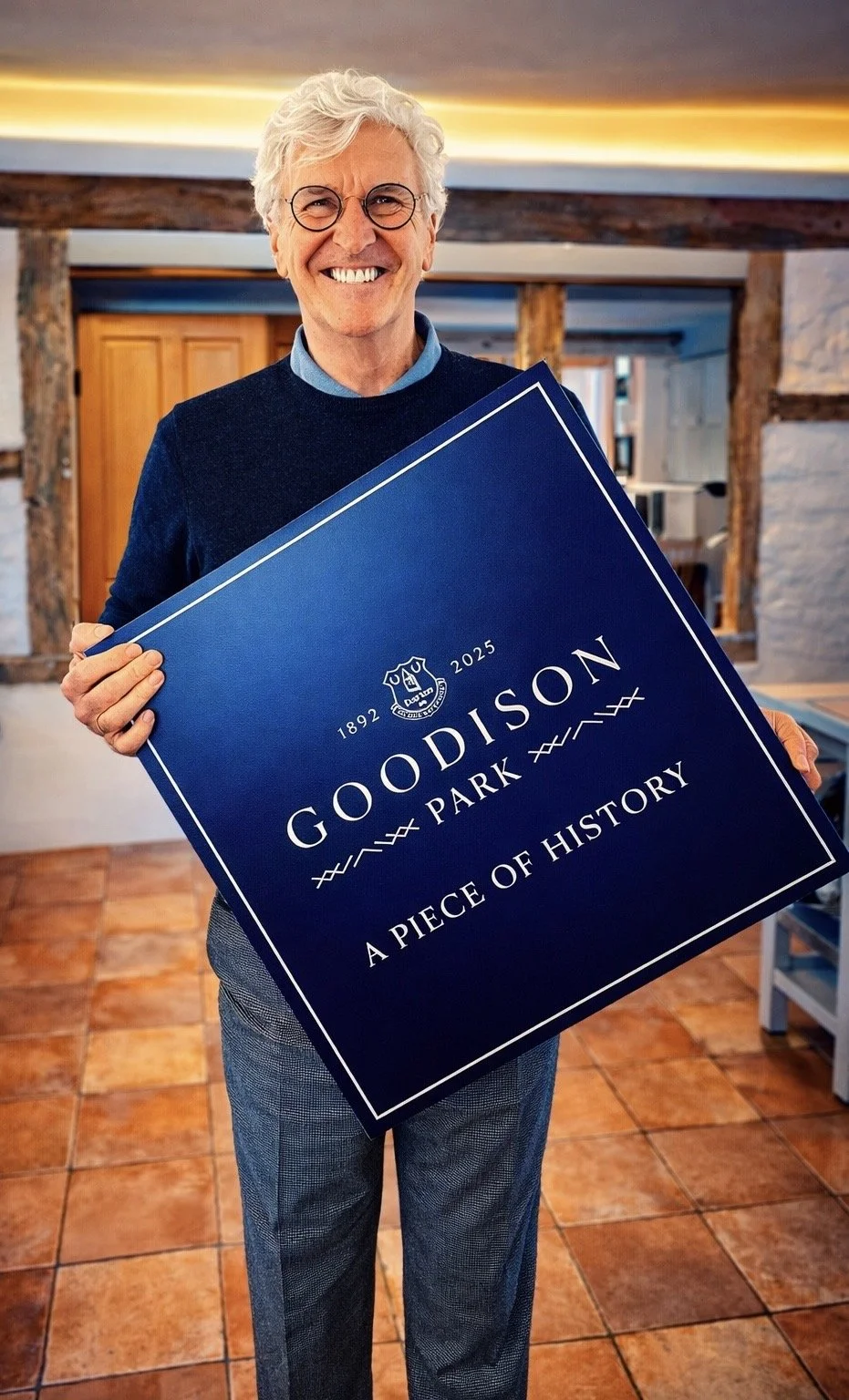 A smiling man with gray hair and round sunglasses, wearing a dark t-shirt, sitting in a modern indoor space with large windows and blurred background.