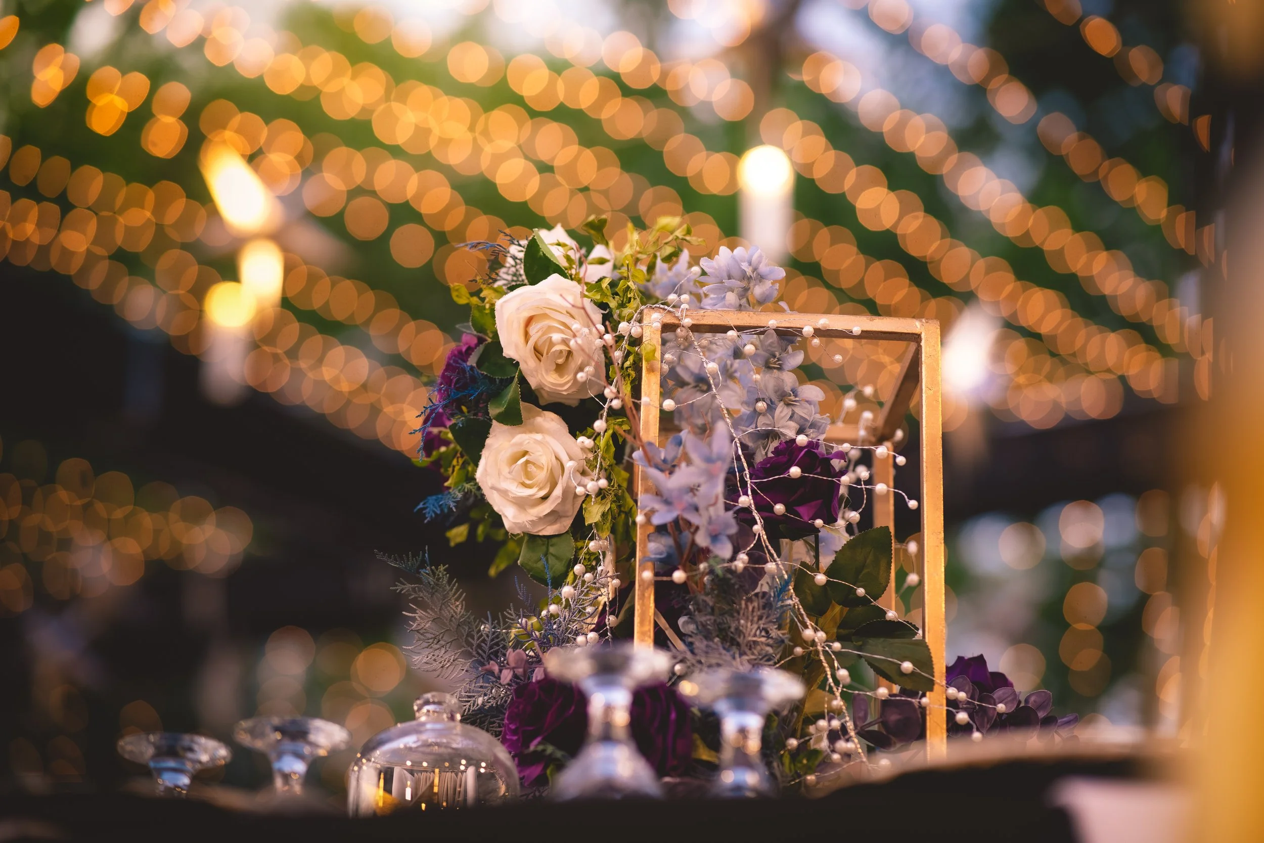 Floral arrangement with white roses, purple flowers, and greenery in a golden geometric frame, surrounded by glassware, with bokeh string lights in the background.
