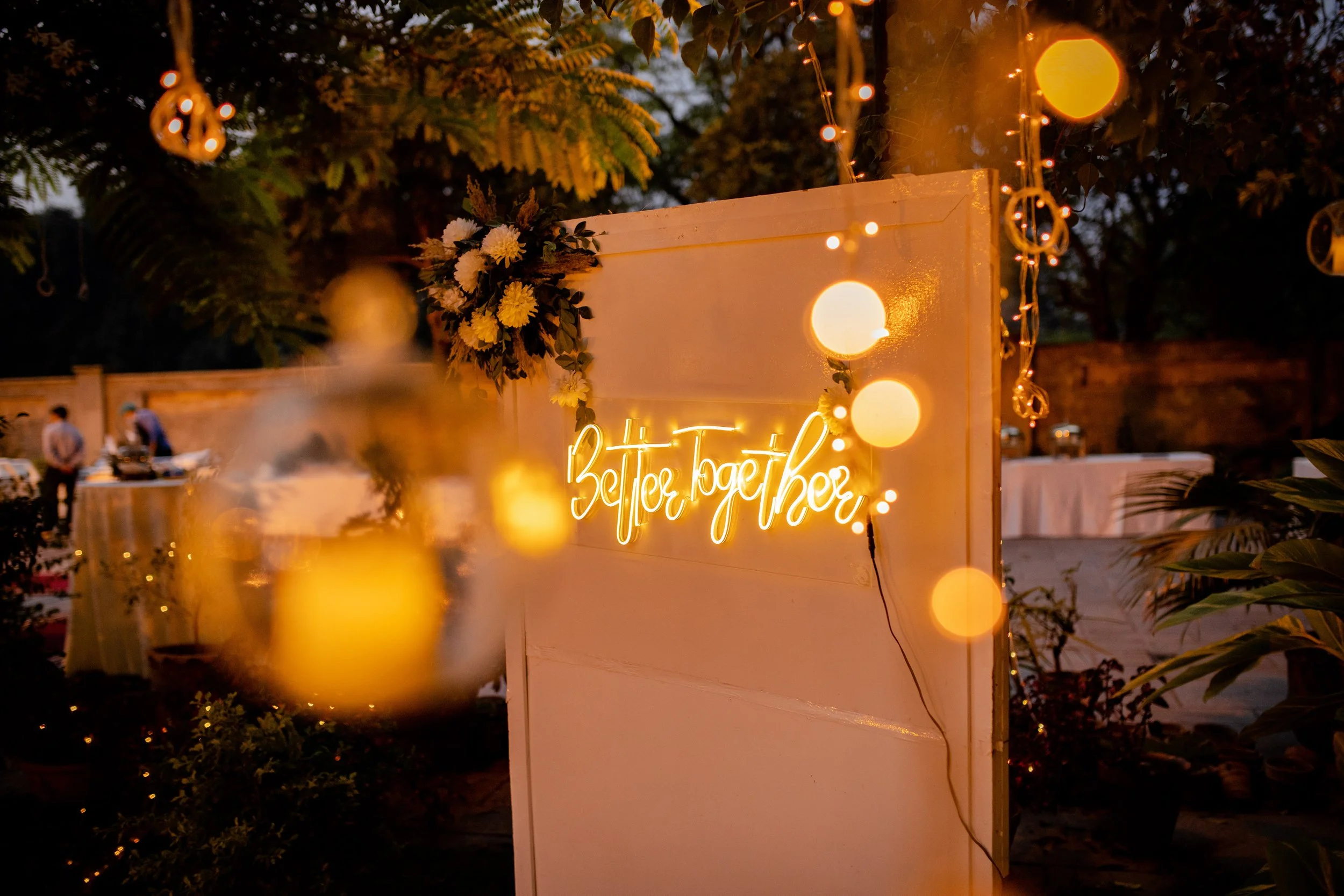 Decorative outdoor event setting with a "Better Together" neon sign, floral arrangement, and string lights.