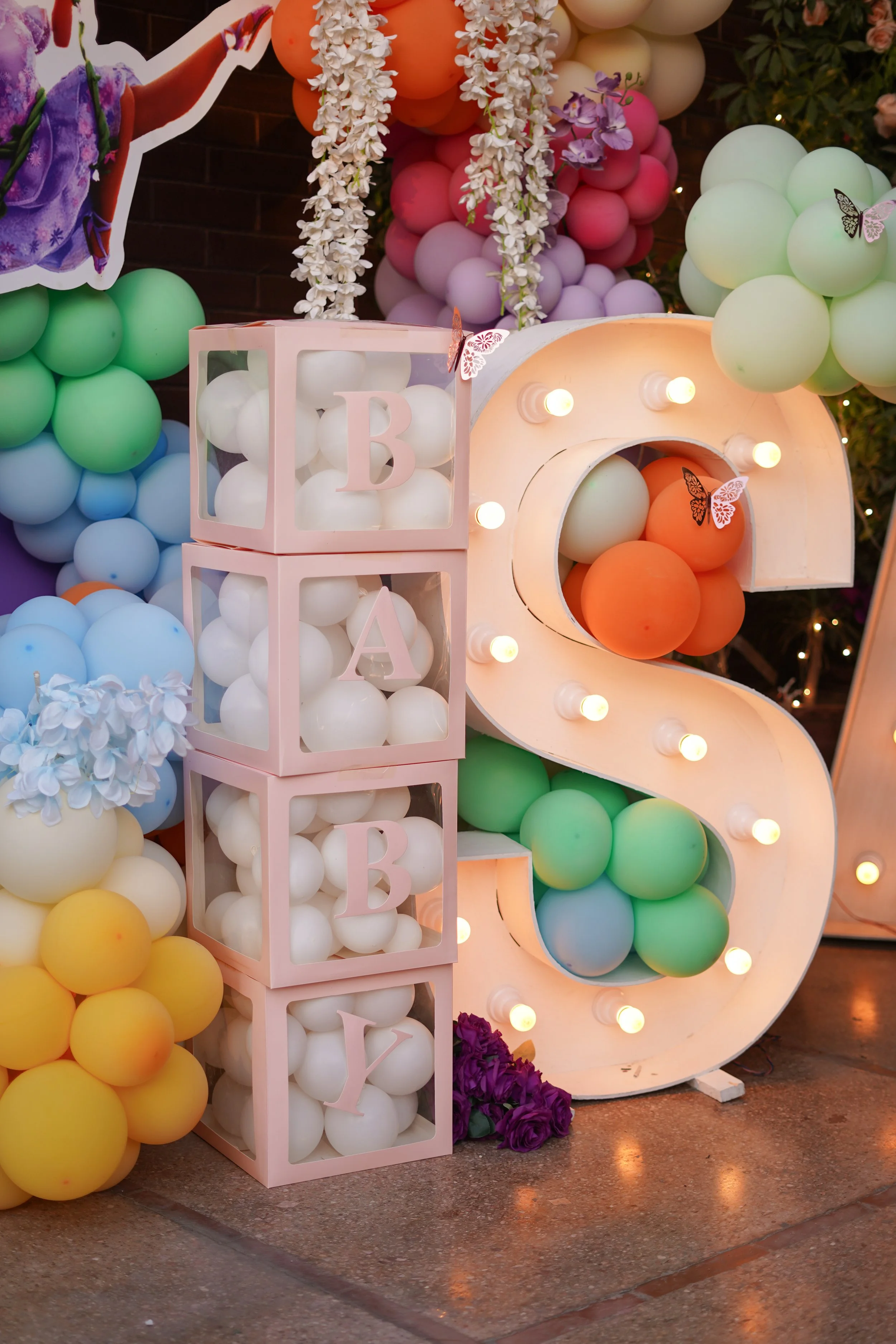 Decorative baby shower display with colorful balloon clusters, illuminated with a large letter "S" and stacked pink boxes spelling "BABY."
