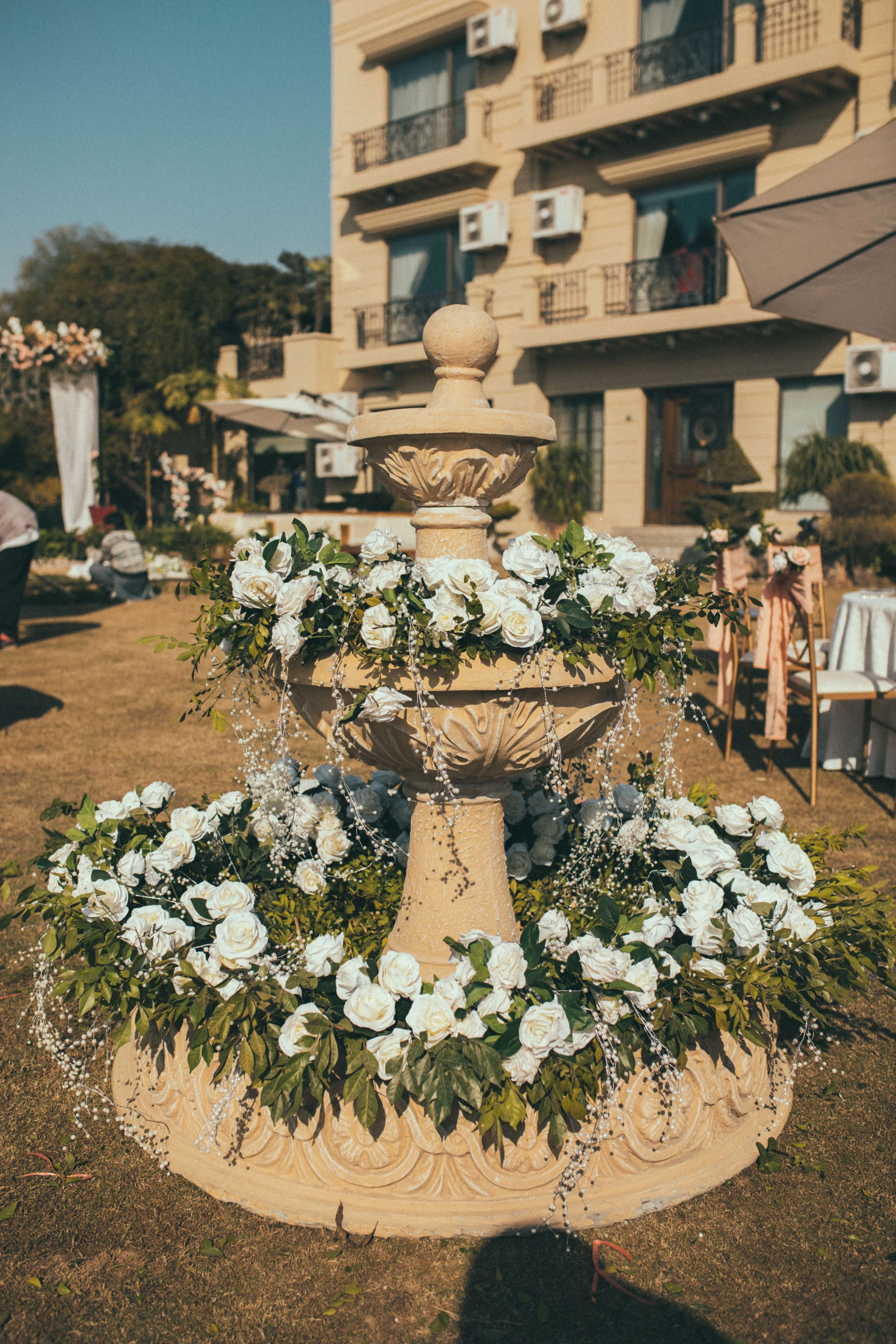 A decorative stone fountain adorned with white roses and greenery in front of a beige building with balconies and air conditioners.
