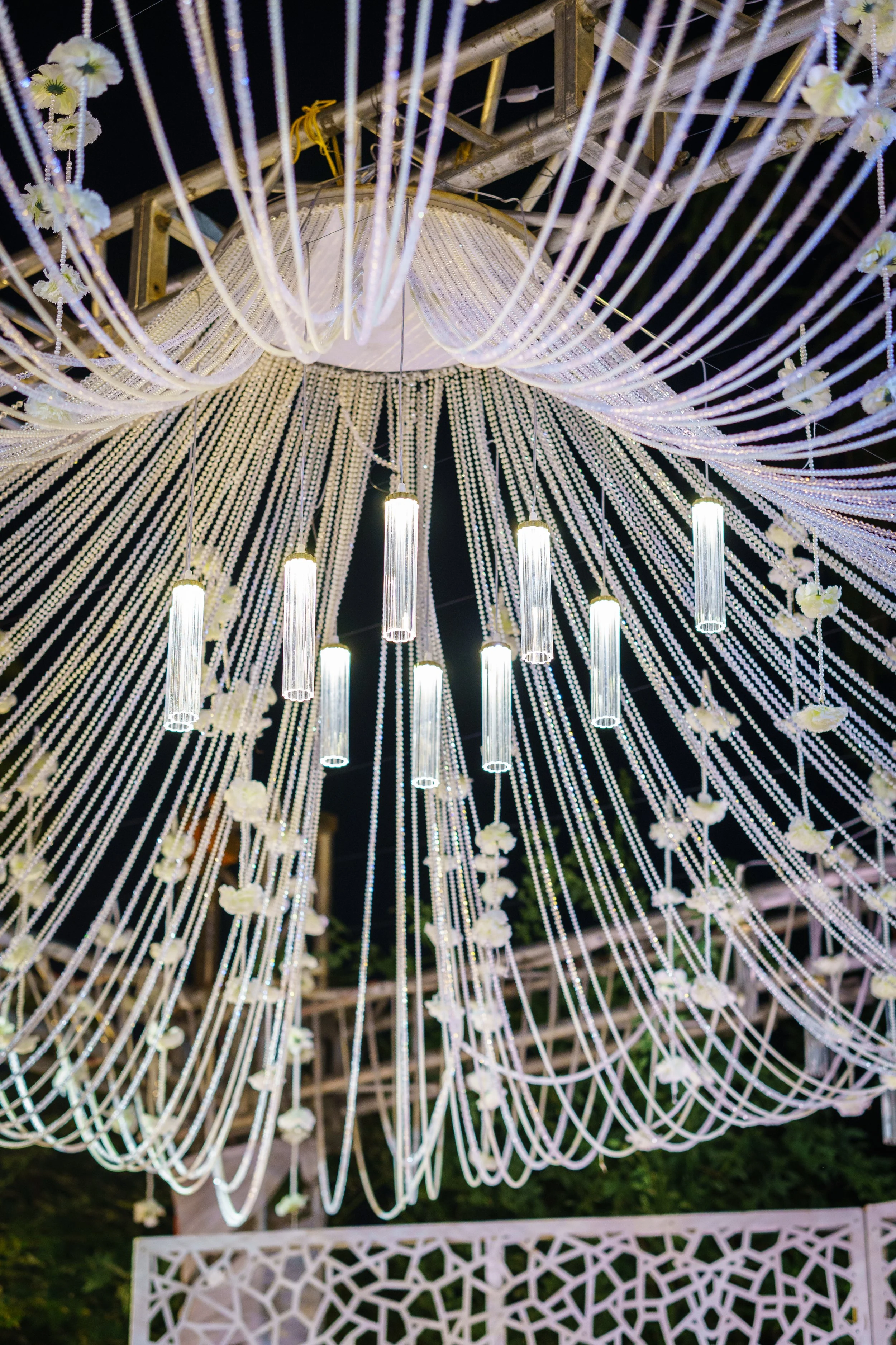 Elaborate wedding decoration with white draped strings, hanging flowers, and illuminated cylindrical lights against a dark background.
