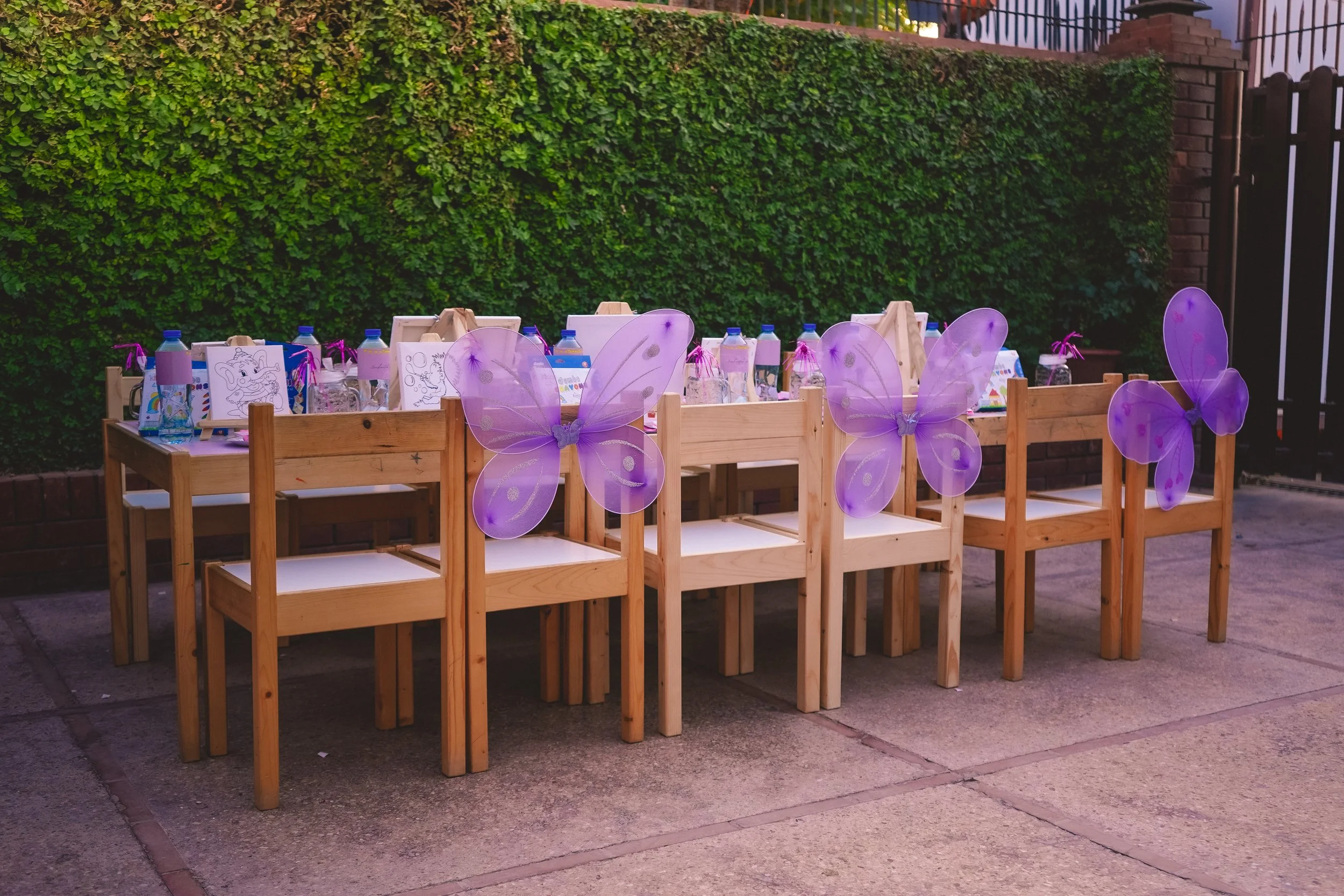 Outdoor party setup with wooden chairs and tables, decorated with purple butterfly wings and covered with drawing materials and drinks, in front of a green ivy wall.