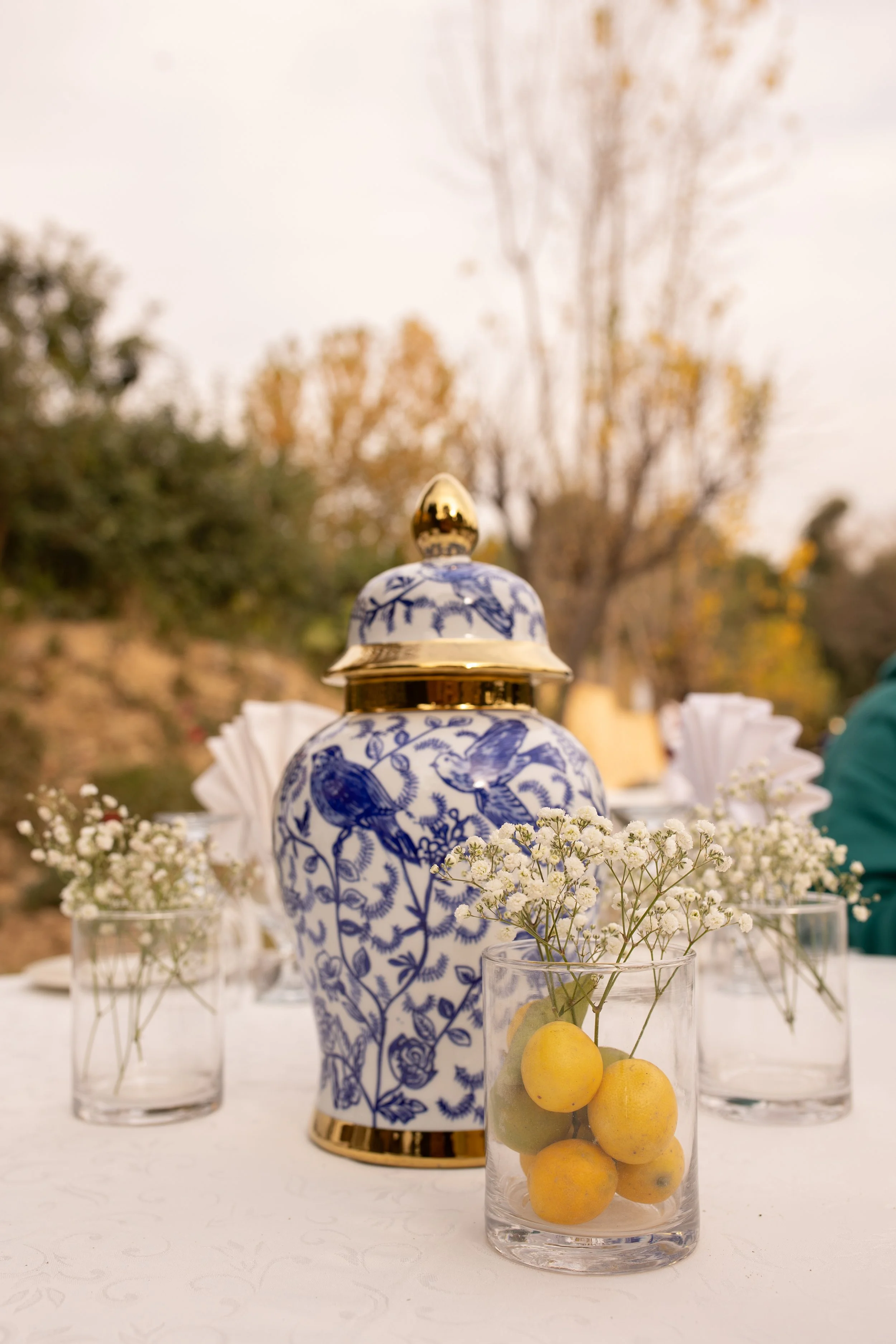 Decorative blue and white ceramic jar with gold accents, surrounded by glass vases holding small white flowers and ripe kumquats, placed on an outdoor table.