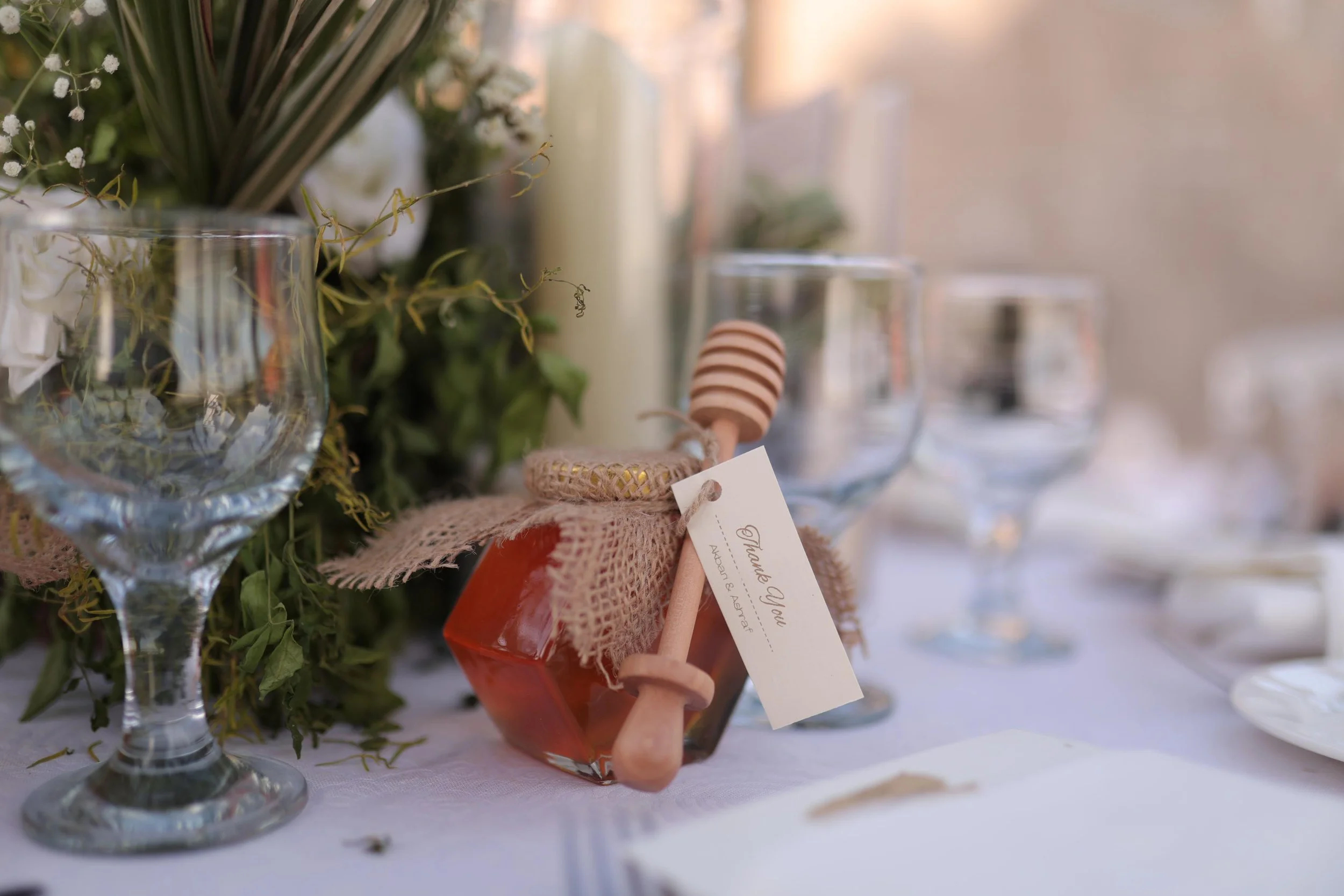 Close-up of a wedding table setting featuring a glass jar of honey with a wooden dipper and thank you tag, surrounded by wine glasses and decorative greenery.