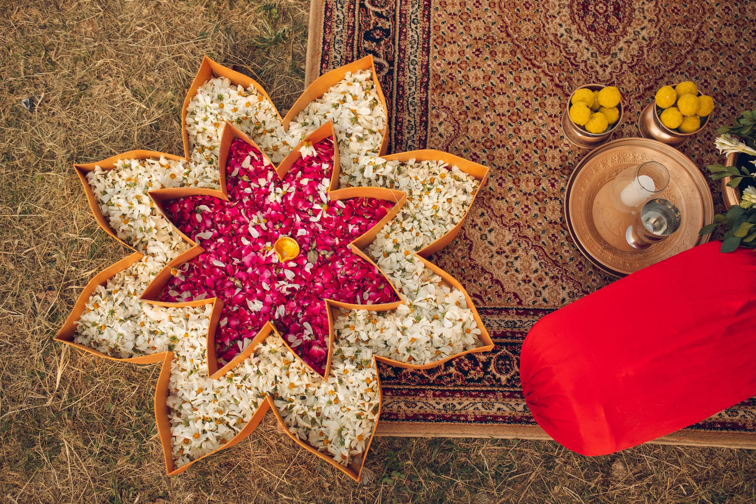 Top view of floral decoration with white and red petals, surrounded by traditional items on grass and a patterned carpet.