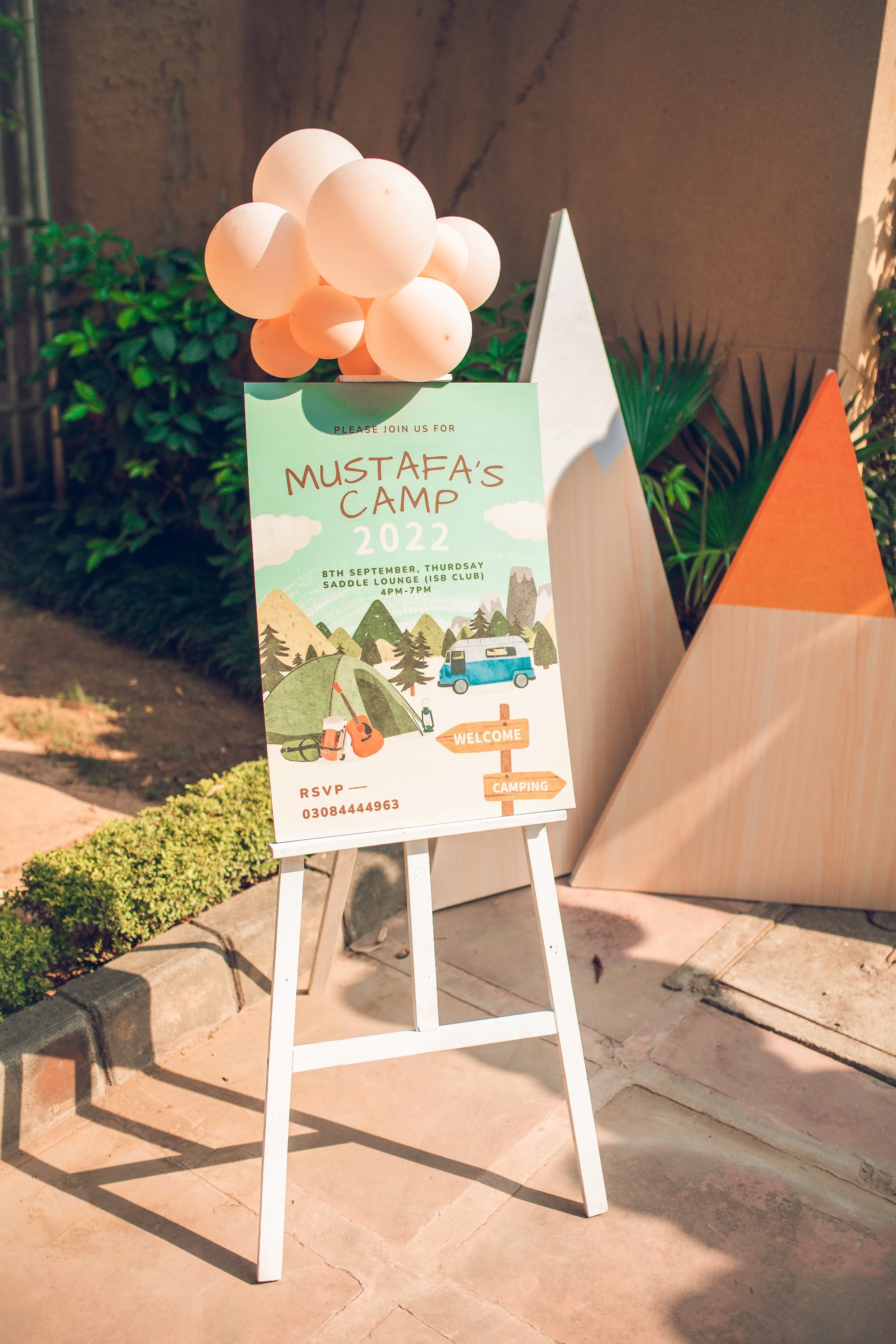 Easel displaying a poster for Mustafa's Camp 2022 with balloons on top, surrounded by plants and triangular decorations.