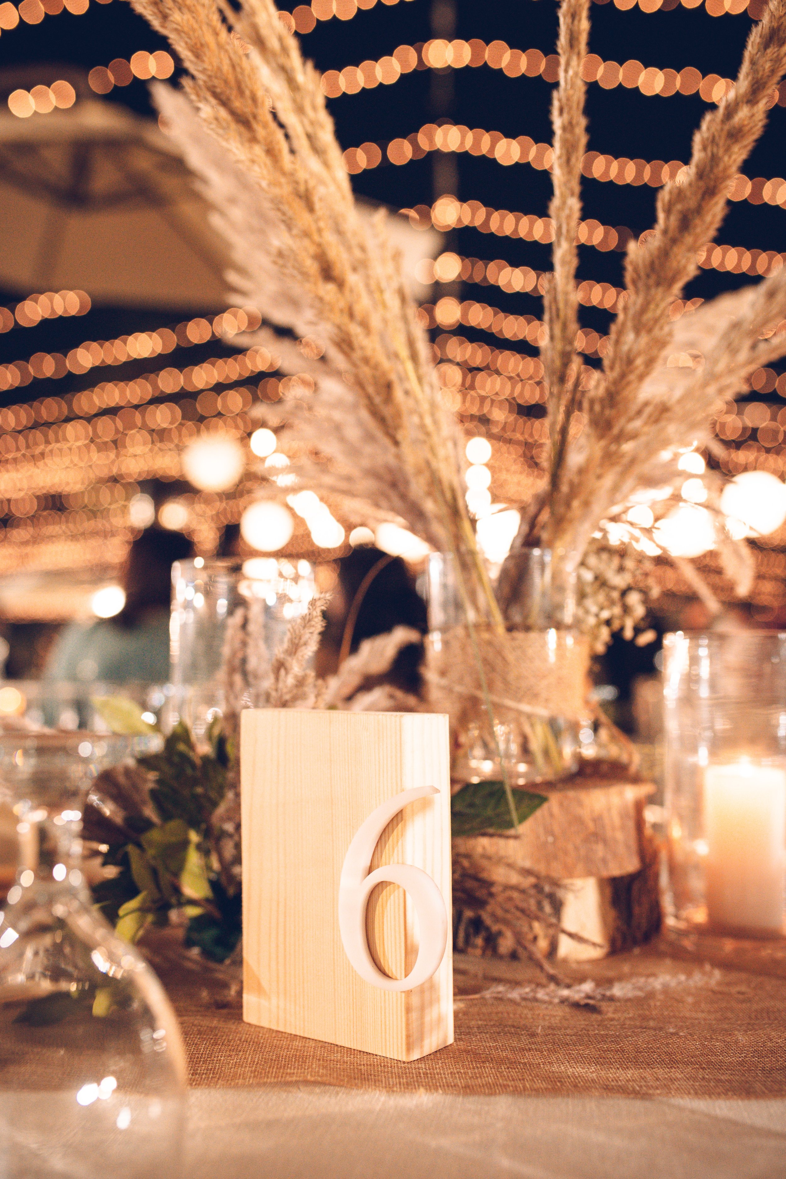 Event table setting with a wooden block displaying number 6, surrounded by decorative pampas grass, candles, and string lights in the background.