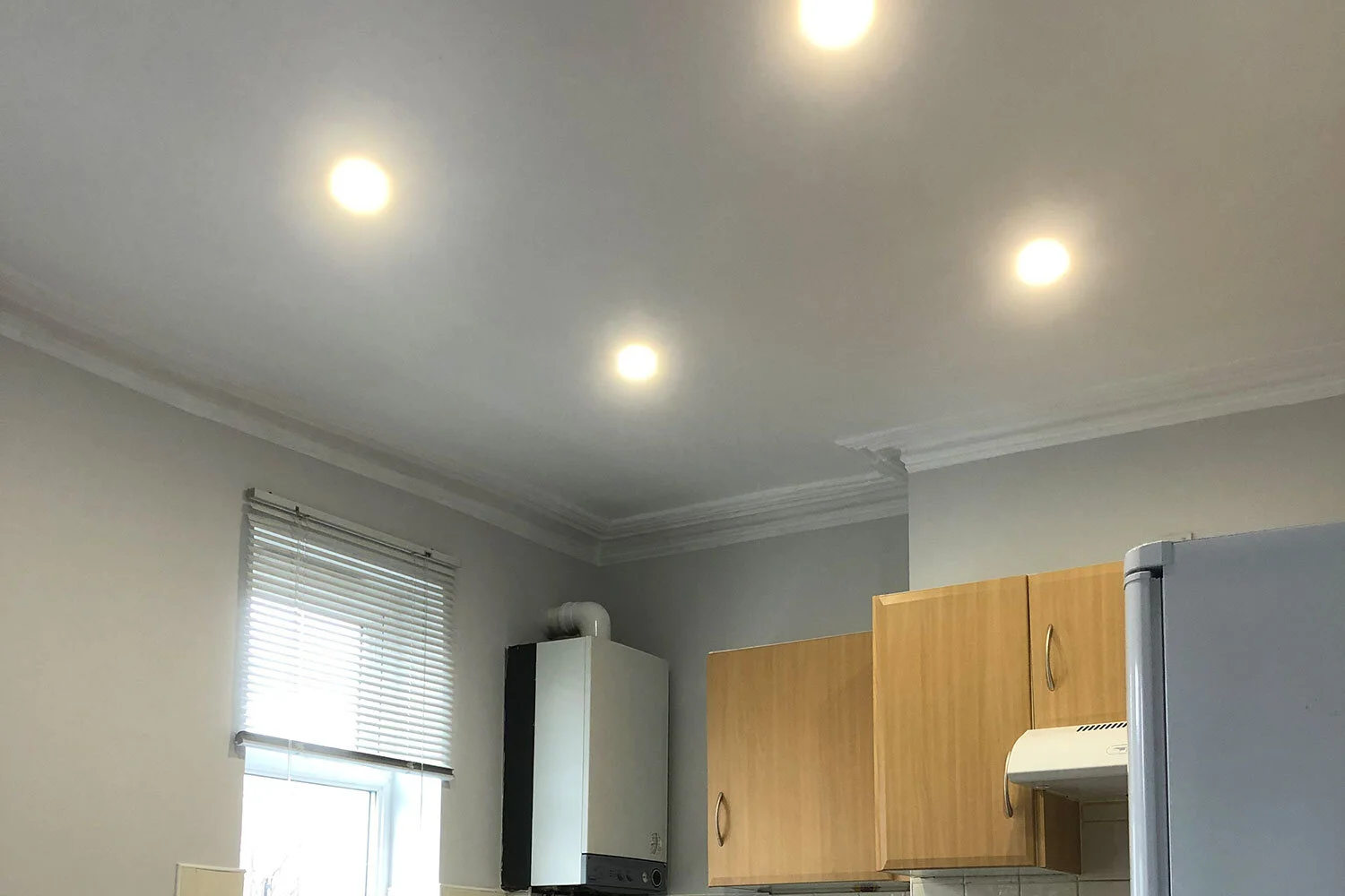 Kitchen ceiling with four recessed lights turned on, gray walls, window with blinds, wooden cabinets.