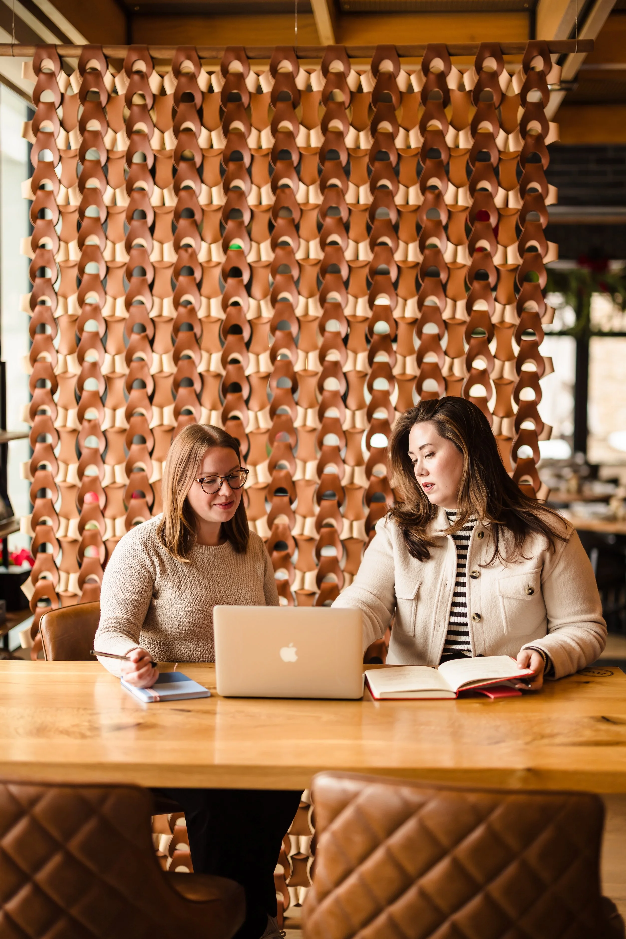 Two women sitting at a wooden table in a restaurant or cafe, engaged in discussion, with a decorative wall of numerous hanging clay or terracotta pots in the background.