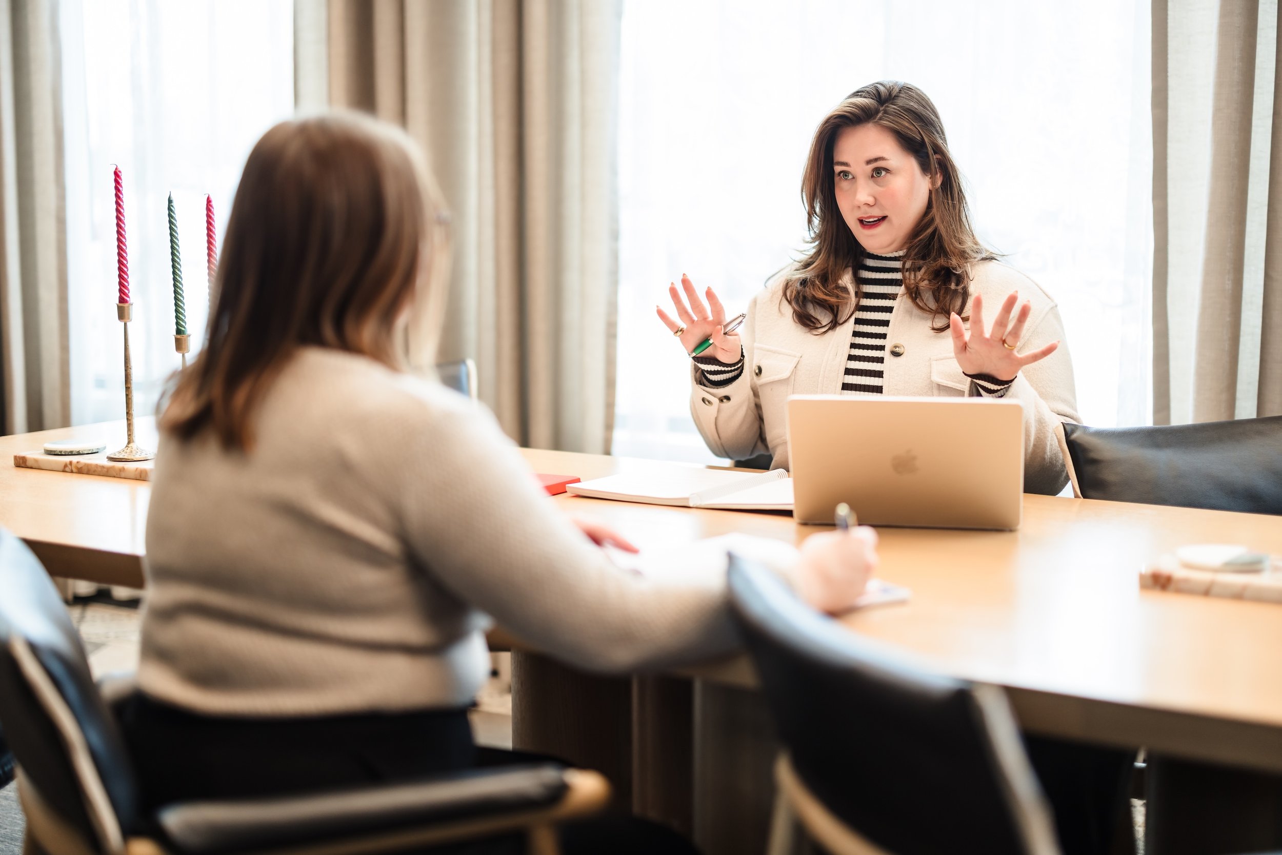 Two women are having a conversation at a table, with one woman speaking and gesturing with her hands, while the other woman listens. There is a laptop and notebooks on the table.
