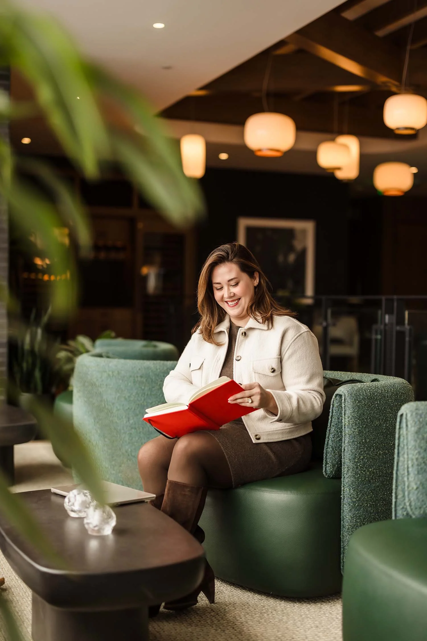 A woman reading a red book while sitting on a green couch in a warmly lit lobby