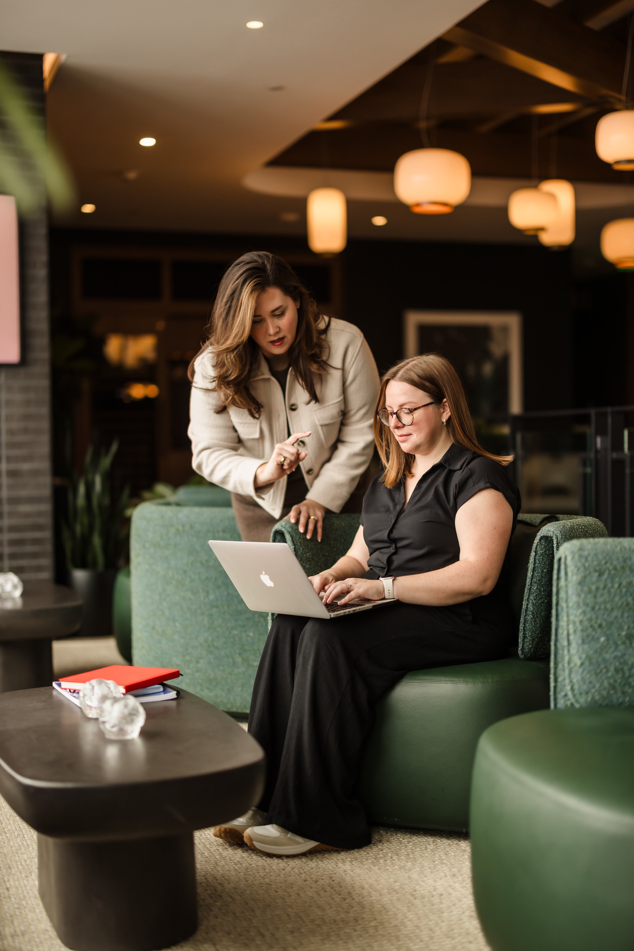 Two women are engaged in a conversation in a modern, warmly lit lobby area. One woman is seated on a green couch with a laptop, while the other woman is standing next to her, pointing at the laptop screen. The seated woman is wearing glasses and a black dress, and the standing woman is wearing a cream-colored jacket.