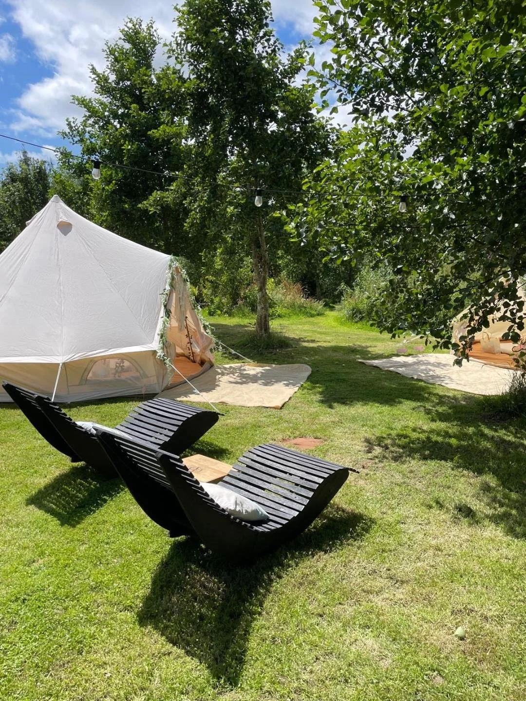 A backyard scene with a white tent, two black wooden lounge chairs with cushions, and an outdoor string light hanging between trees on a sunny day.
