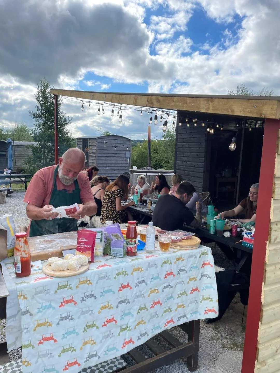 People gathered outdoors for a barbecue or picnic, with a man preparing food in the foreground and others seated at a long table with food and drinks under a wooden shelter with string lights. The sky is partly cloudy.