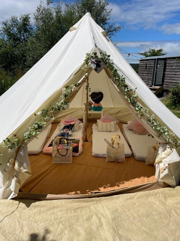 Inside a white canvas tent decorated with greenery, with cushions and small beds arranged for resting or sleeping, decorated with pillows and face masks. There are also shopping bags in the tent, and it is set outdoors on a sunny day with trees and a