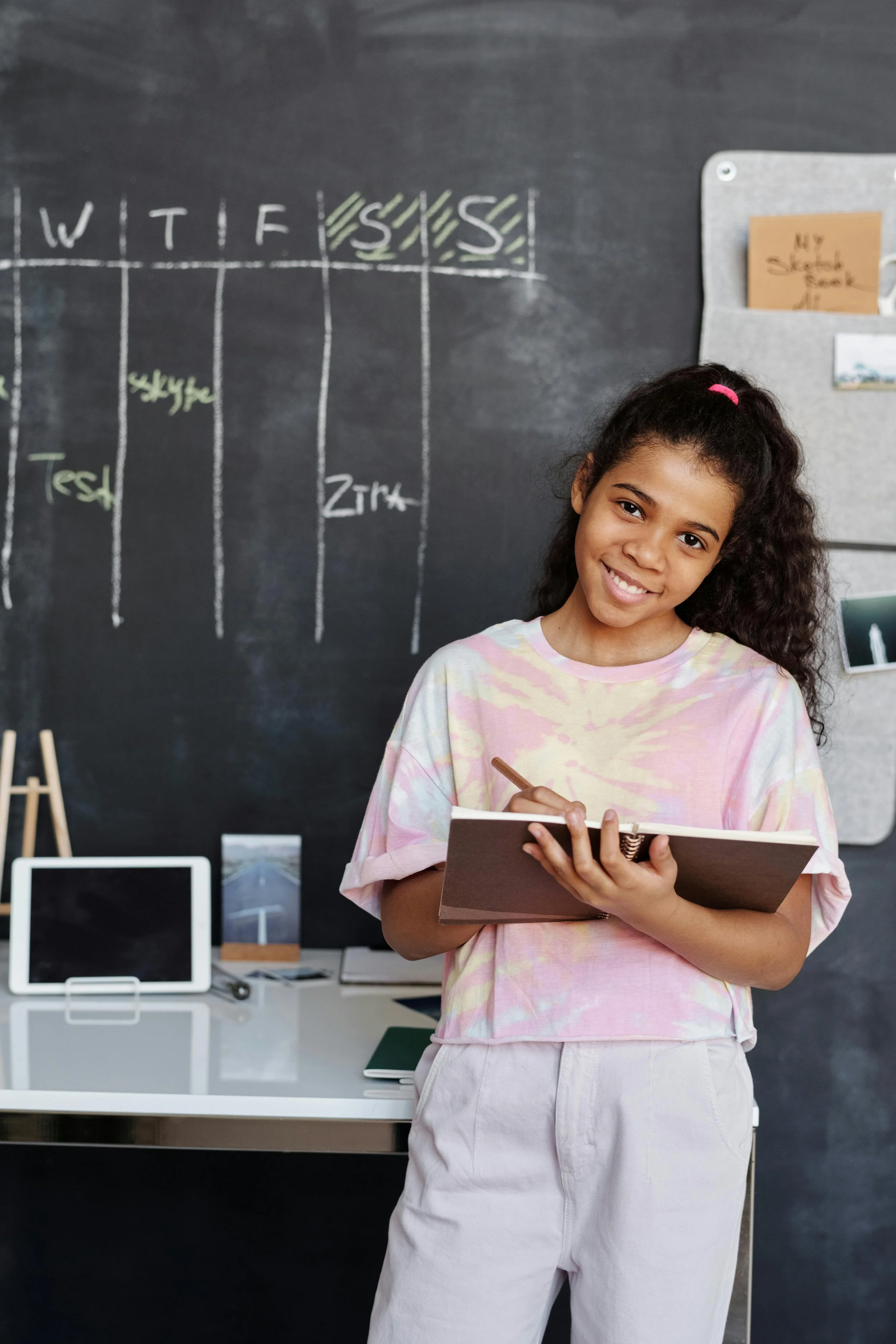 A young girl with curly hair, wearing a pink tie-dye shirt and light pants, is smiling and holding a notebook and pen in a classroom with a wall chalkboard and a desk with a tablet and books.