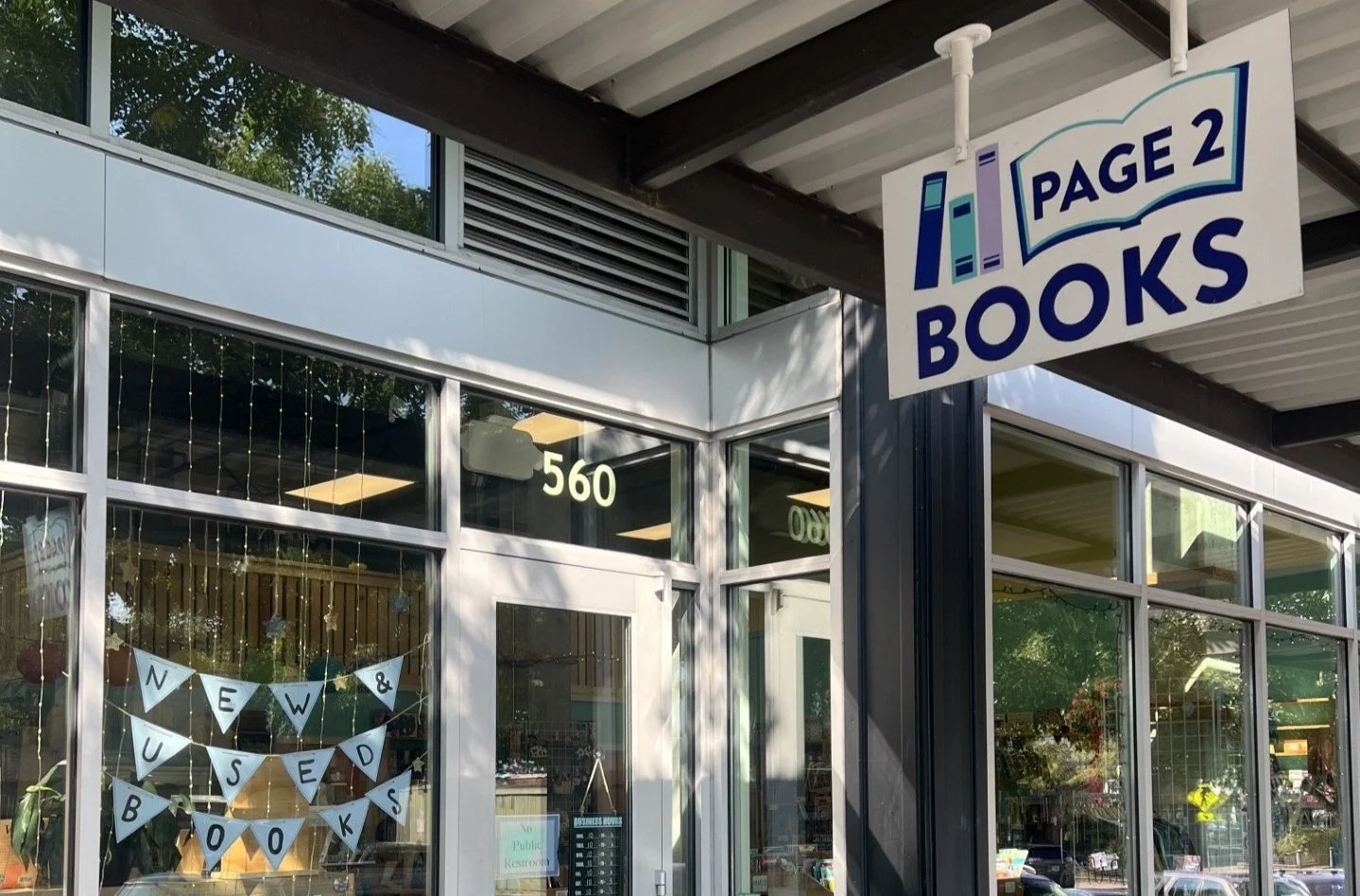 A photo of the outside of the store. The window has string lights and a triangle banner saying "New & Used Books". Hanging above the door is the store logo which says "Page 2 Books" in dark blue with blue and purple books.
