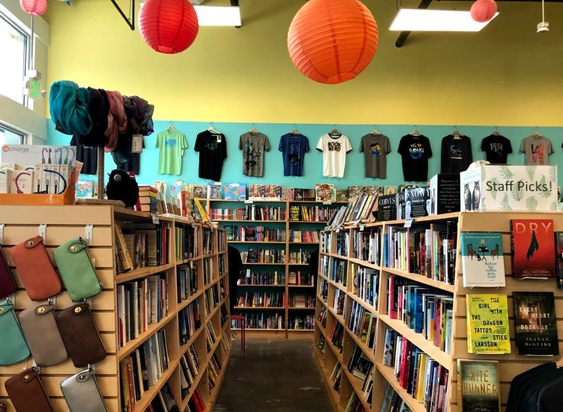 A photo of the inside of the bookstore, showing filled light wood bookshelves and tshirts hung up high on the wall above the shelves.