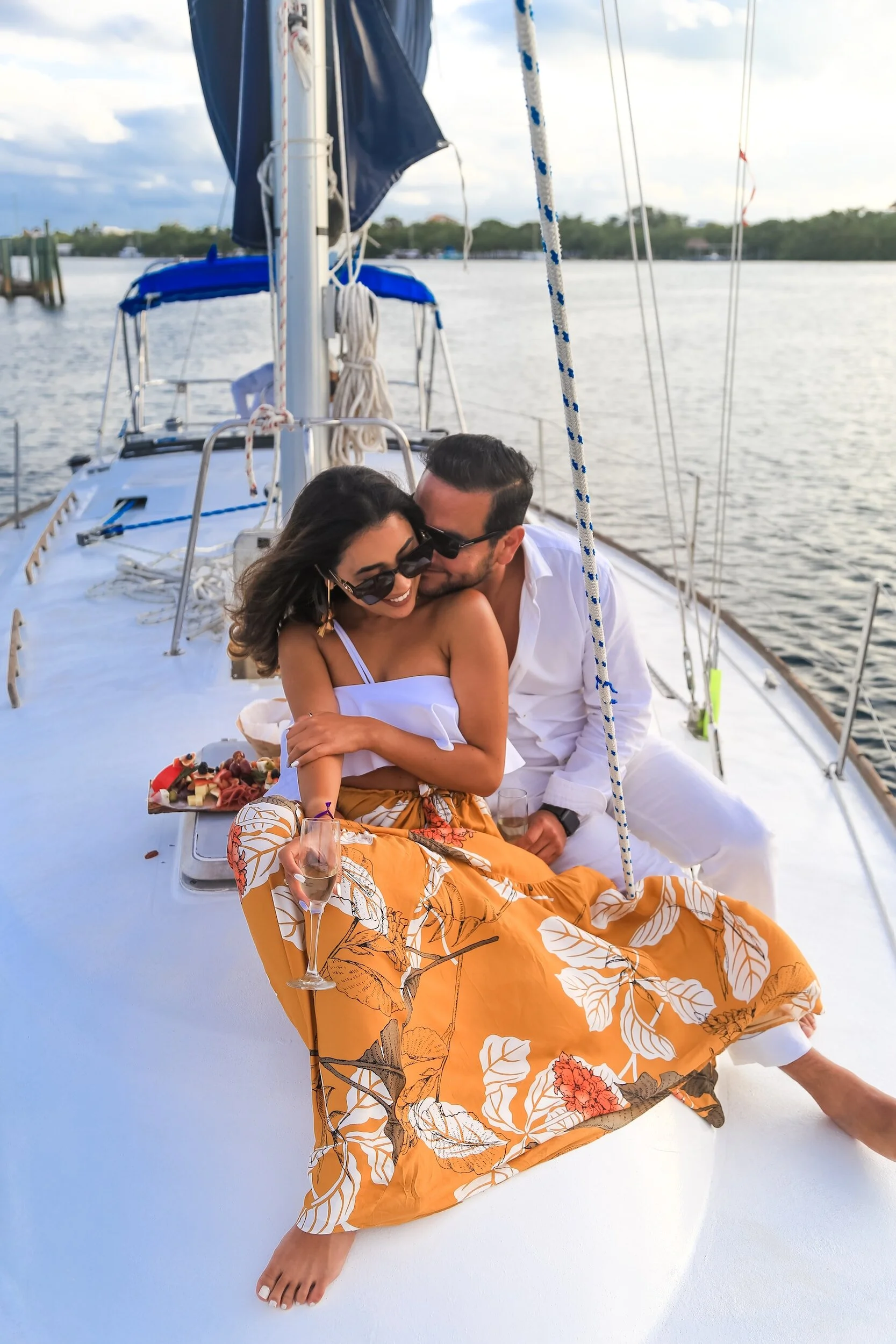 A couple sitting on a boat, smiling and enjoying drinks, with a woman in a white top and orange skirt and a man in a white outfit, surrounded by water and sky.