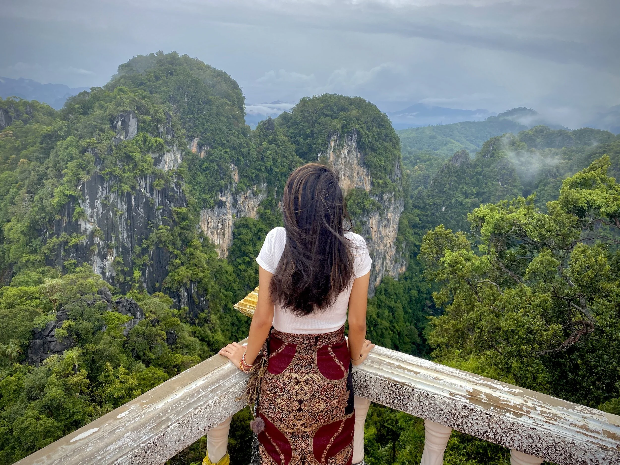 A woman with long dark hair wearing a white shirt and a patterned red and gold skirt is sitting on a wooden railing, looking out at lush green mountains and dense forest under a cloudy sky.