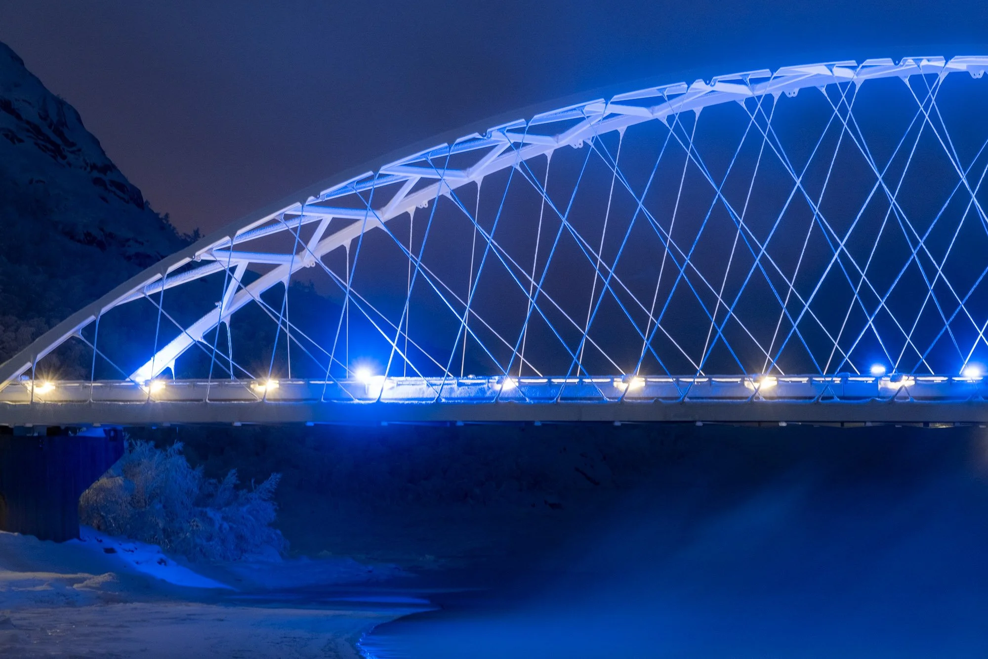 Neonbelyst, modern bro over en frossen elv under en natt med mørk himmel og fjell i bakgrunnen.