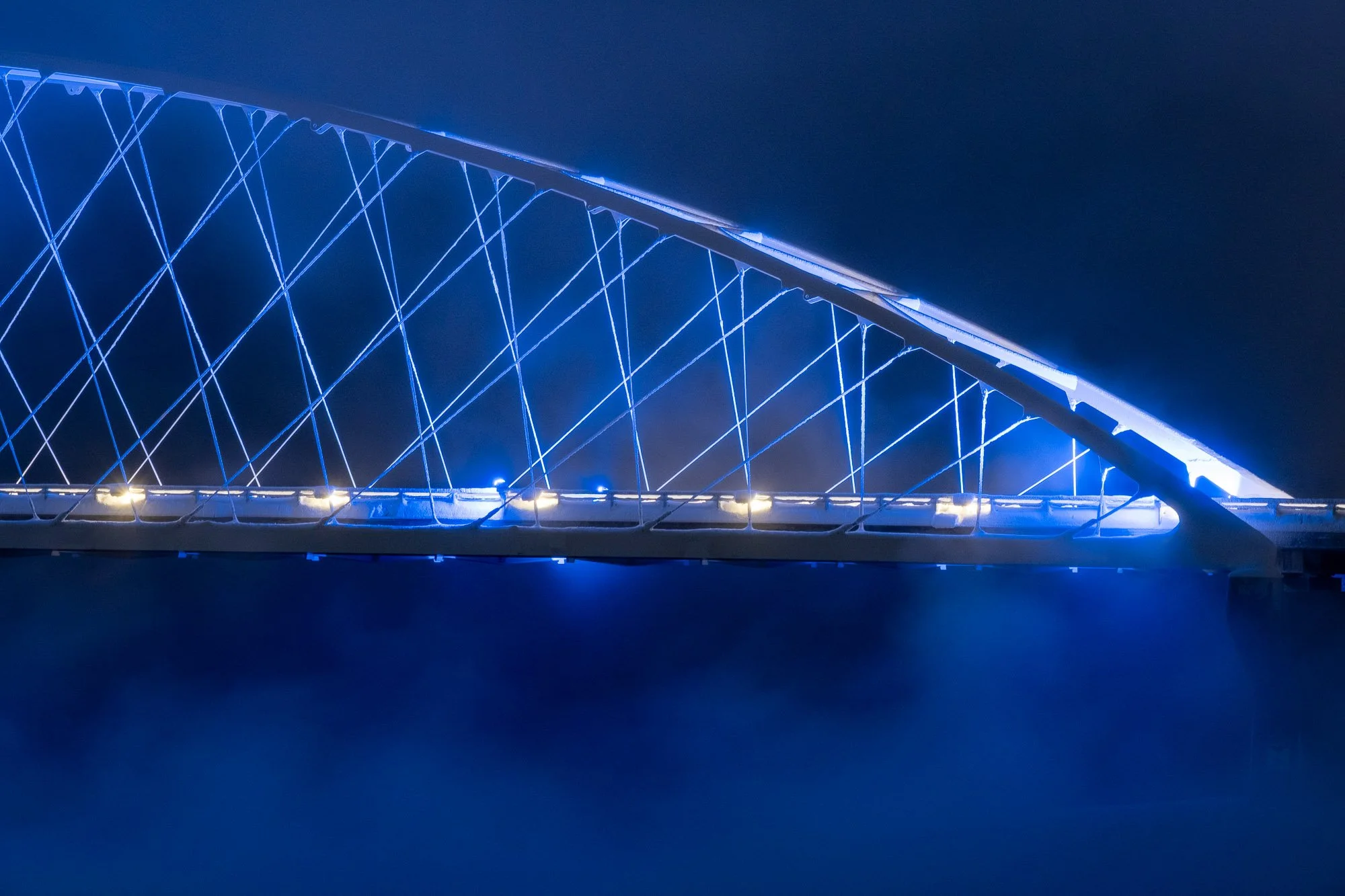 Nattbilde av en pedestrianbro med blå LED-belysning over en vannflate.