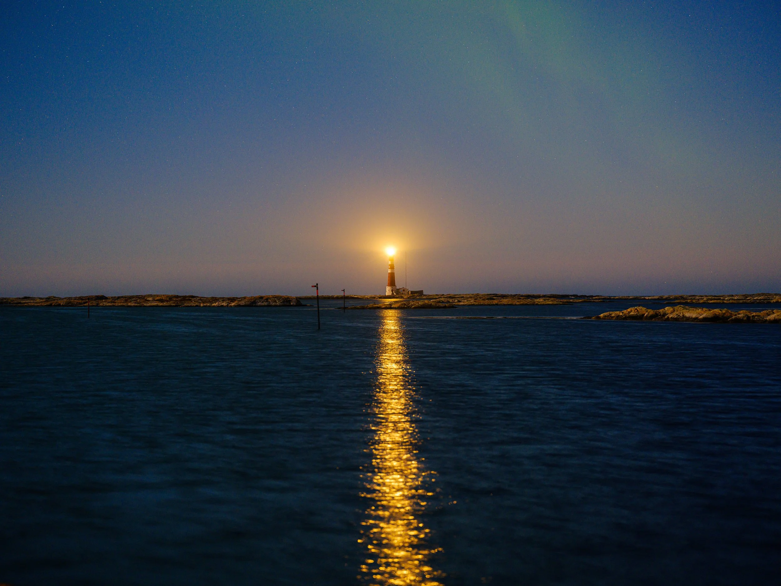 En fyrtårn står på en øy om natten, med lys som lyser opp himmelen og reflekteres i vannet foran.