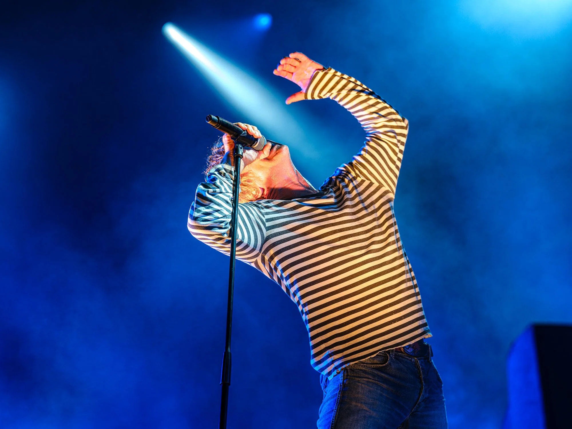 En mann med stripete genser som synger inn i en mikrofon på en scene med blå og hvite lys.