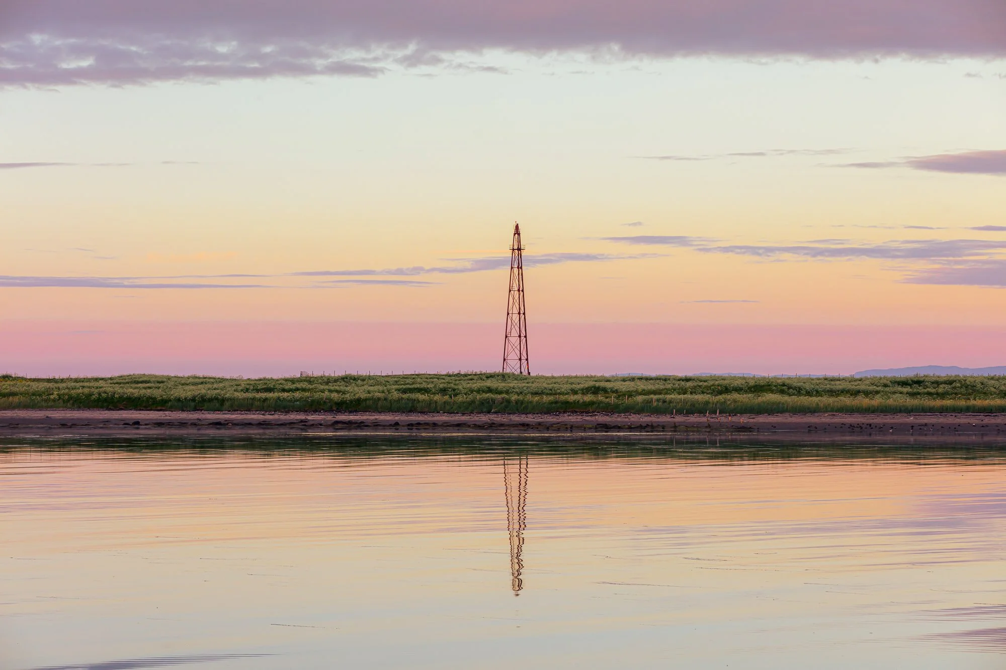 Fott av vann med en ås og en jernbanetårn i bakgrunnen ved solnedgang, fargerik himmel med spredte skyer