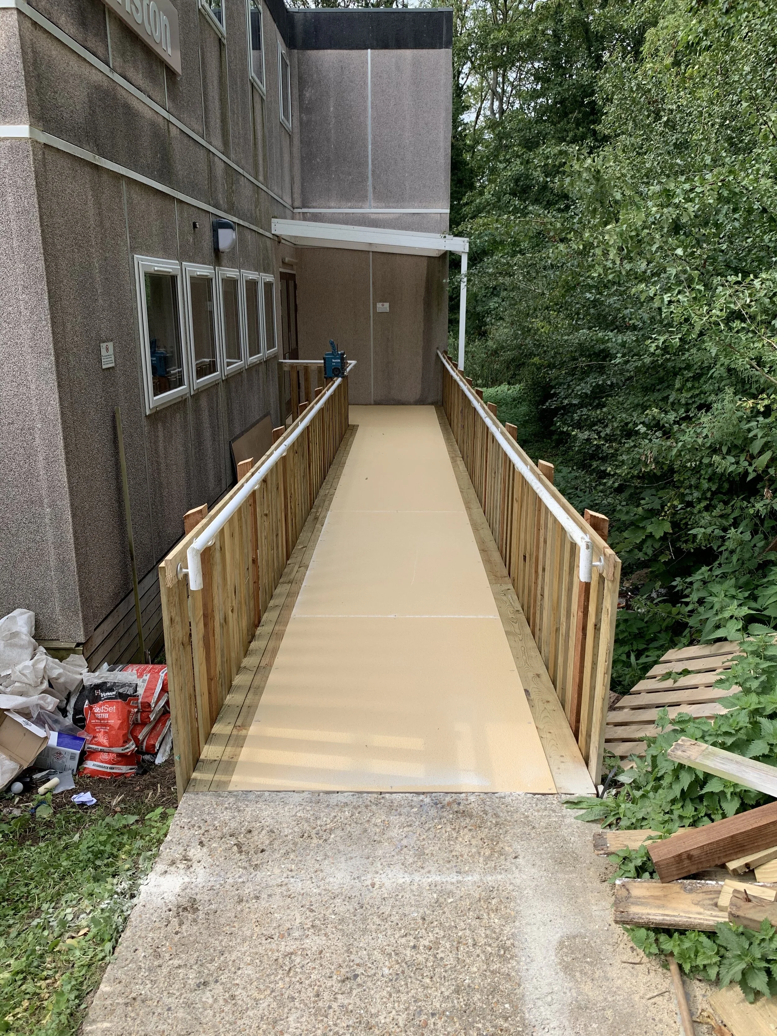 A newly constructed wheelchair accessible ramp leading to an entrance of a building, with wood railings on both sides and some construction materials and debris nearby.