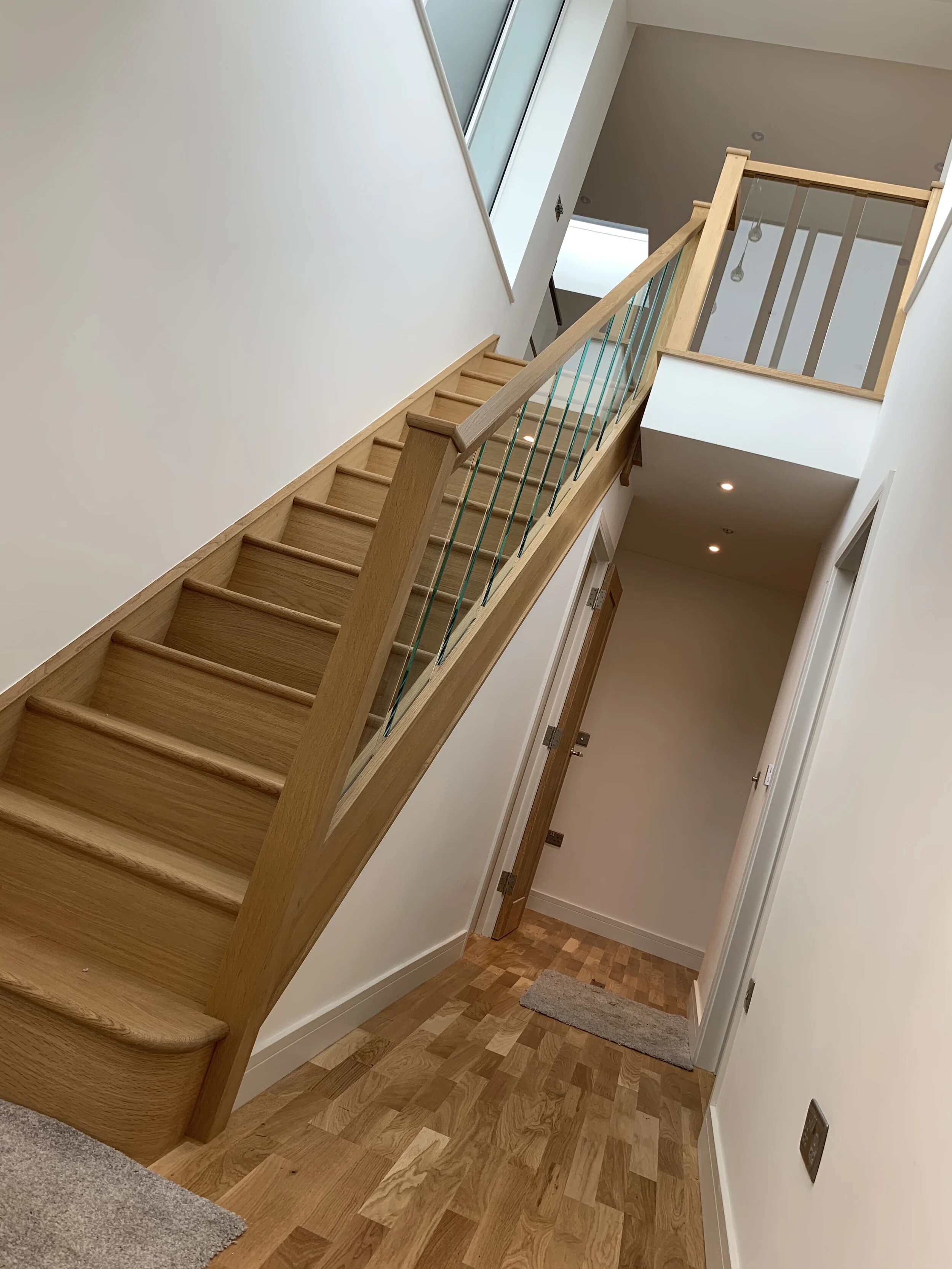 Interior of a modern house showing a wooden staircase with a simple railing, viewed from the ground floor towards the upper floor, with wooden flooring and white walls.