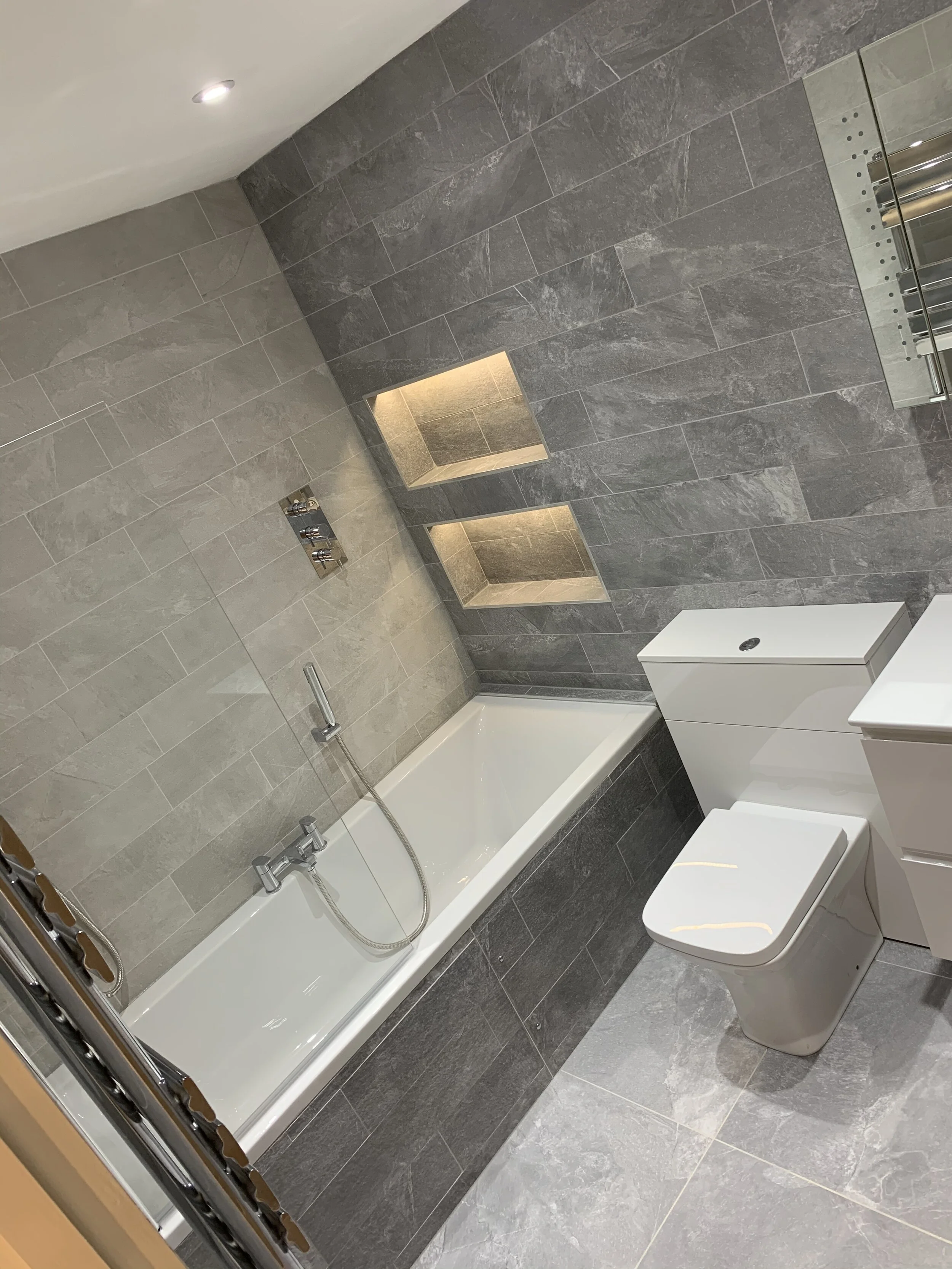 Modern bathroom with gray tiled walls and floor, a built-in shelf with lighting, a white bathtub with a handheld shower, a white vanity with a sink, and a toilet.