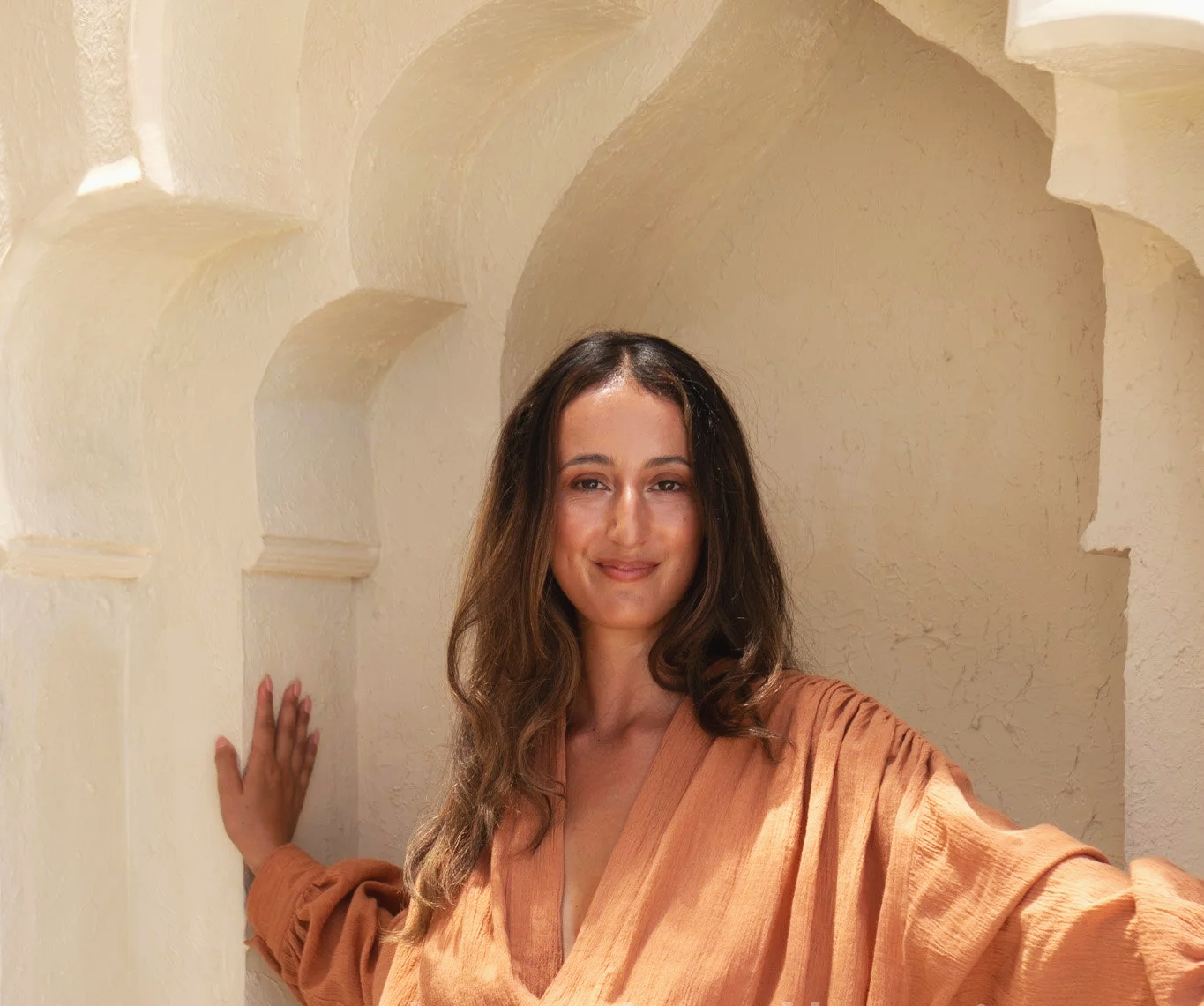 A woman with long wavy brown hair and light skin smiling, wearing a loose orange-brown dress, standing next to a textured beige wall with arched niches.
