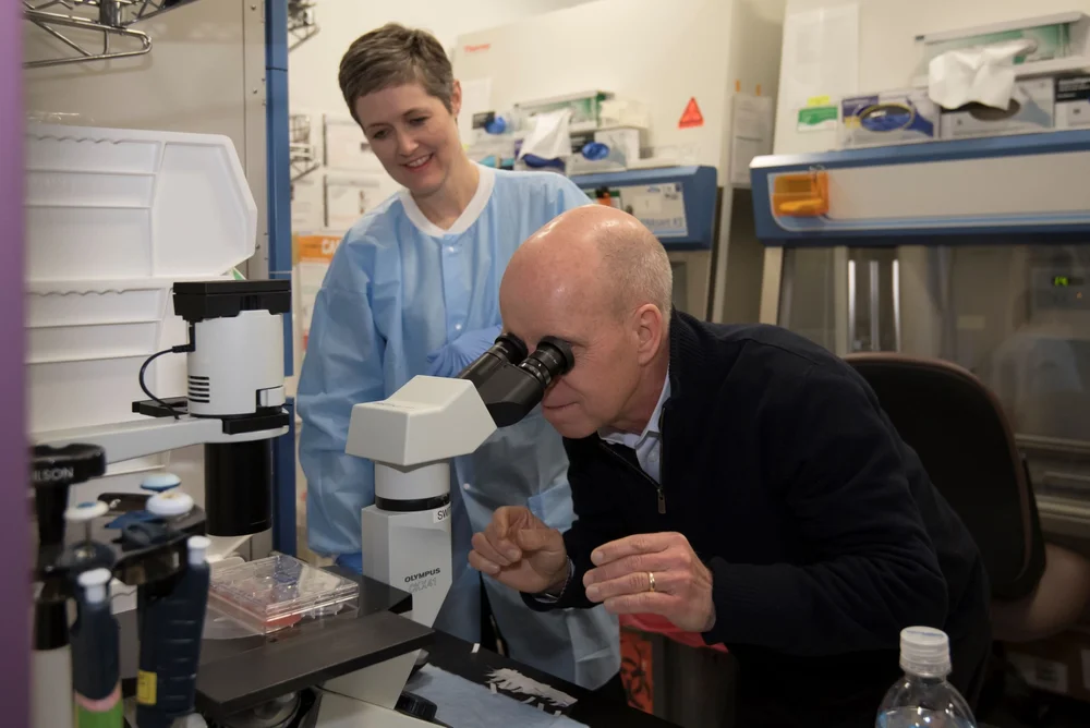 Scott Hamilton looks through a microscope in a cancer research lab as a researcher in a blue lab gown looks on, showing the research his foundation funds