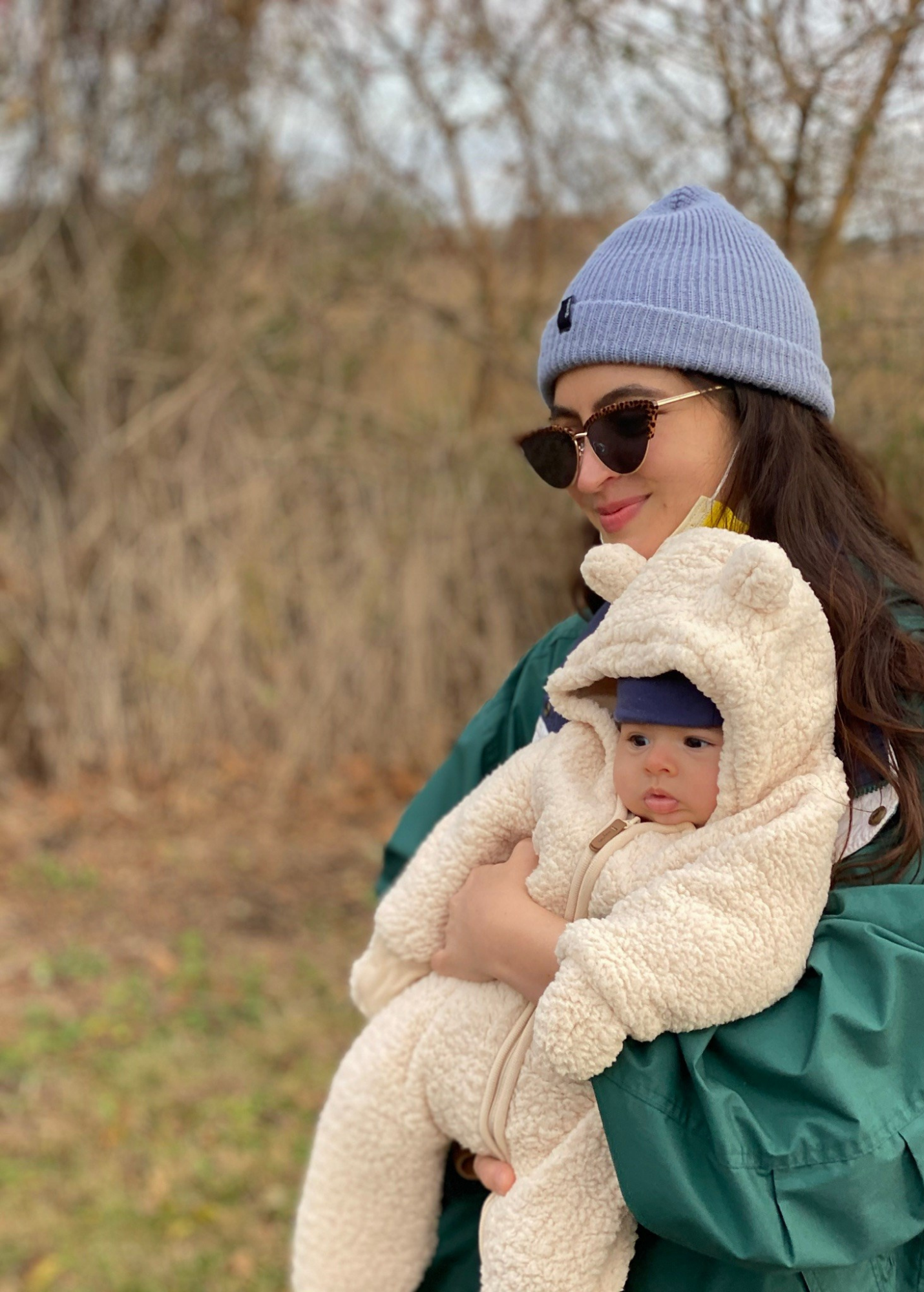 A woman wearing sunglasses, a blue beanie, and a green jacket holds a baby dressed in a beige fluffy jacket and a navy hat outdoors in a park with leafless trees in the background.