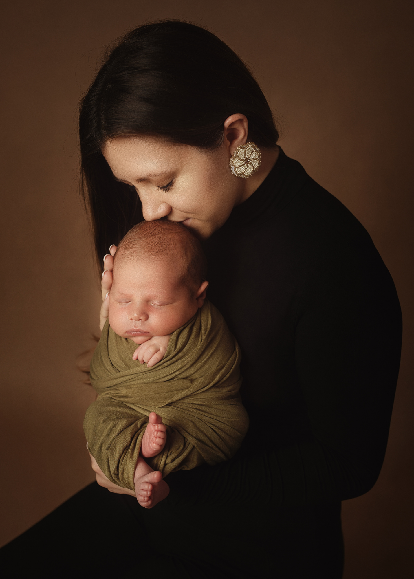 A woman with dark hair, wearing large, ornate earrings, holds a sleeping baby wrapped in a green blanket against a plain brown background.