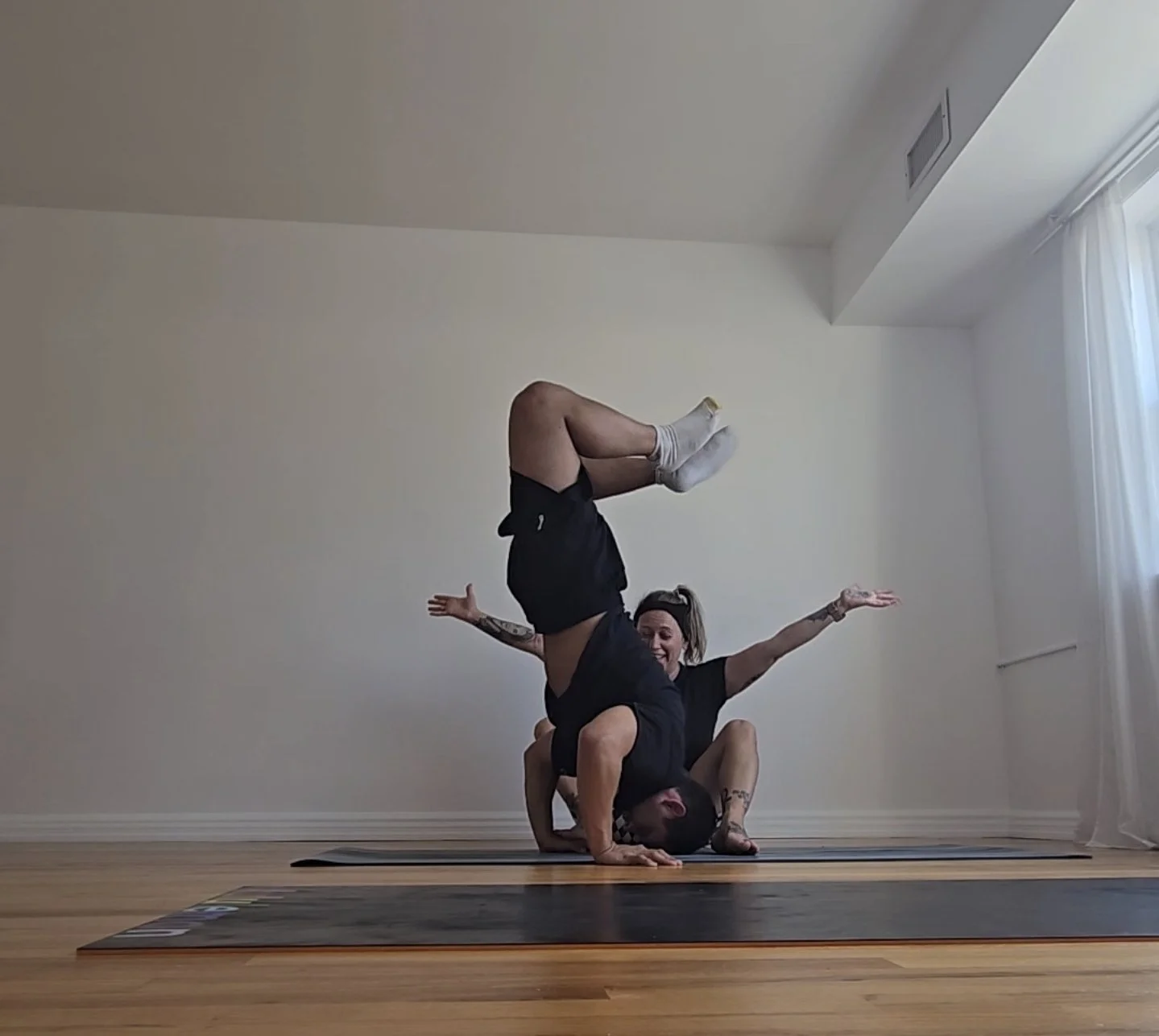 Two women practicing acro yoga in a spacious, well-lit room with wooden floors, one balancing upside down on the other's shoulders, both smiling, with arms outstretched.