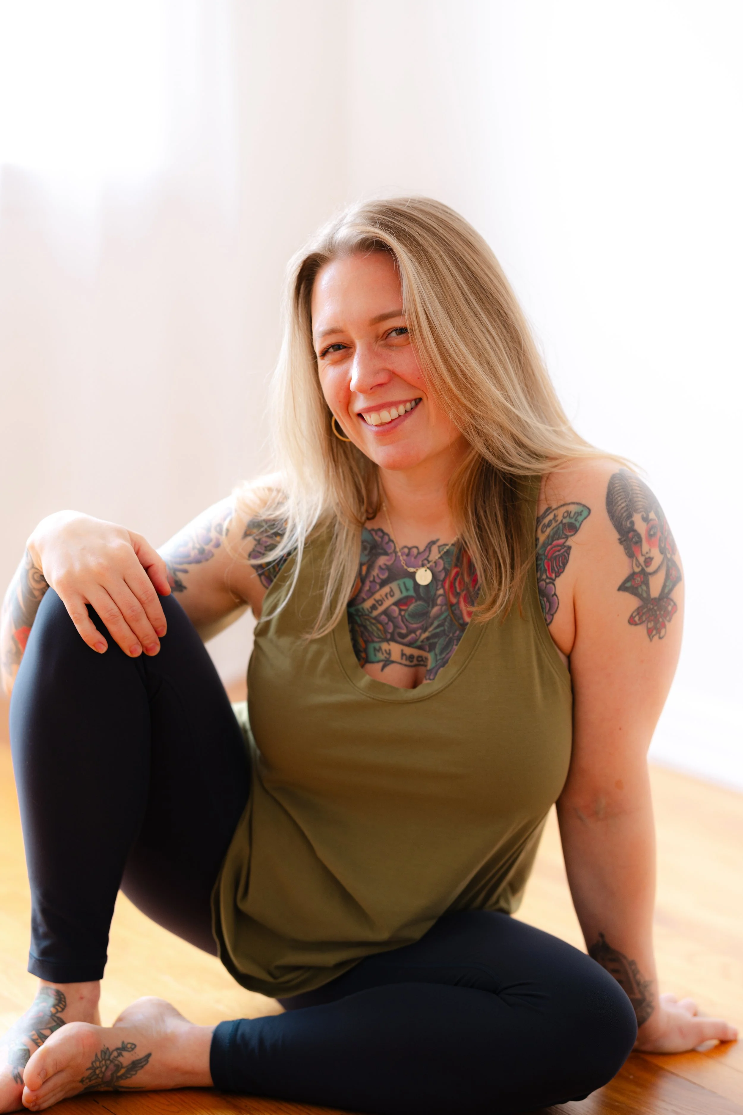 A woman with blonde hair and tattoos on her arms and neck, sitting in a yoga pose on a wooden floor with a bright, neutral background, smiling while looking at the camera