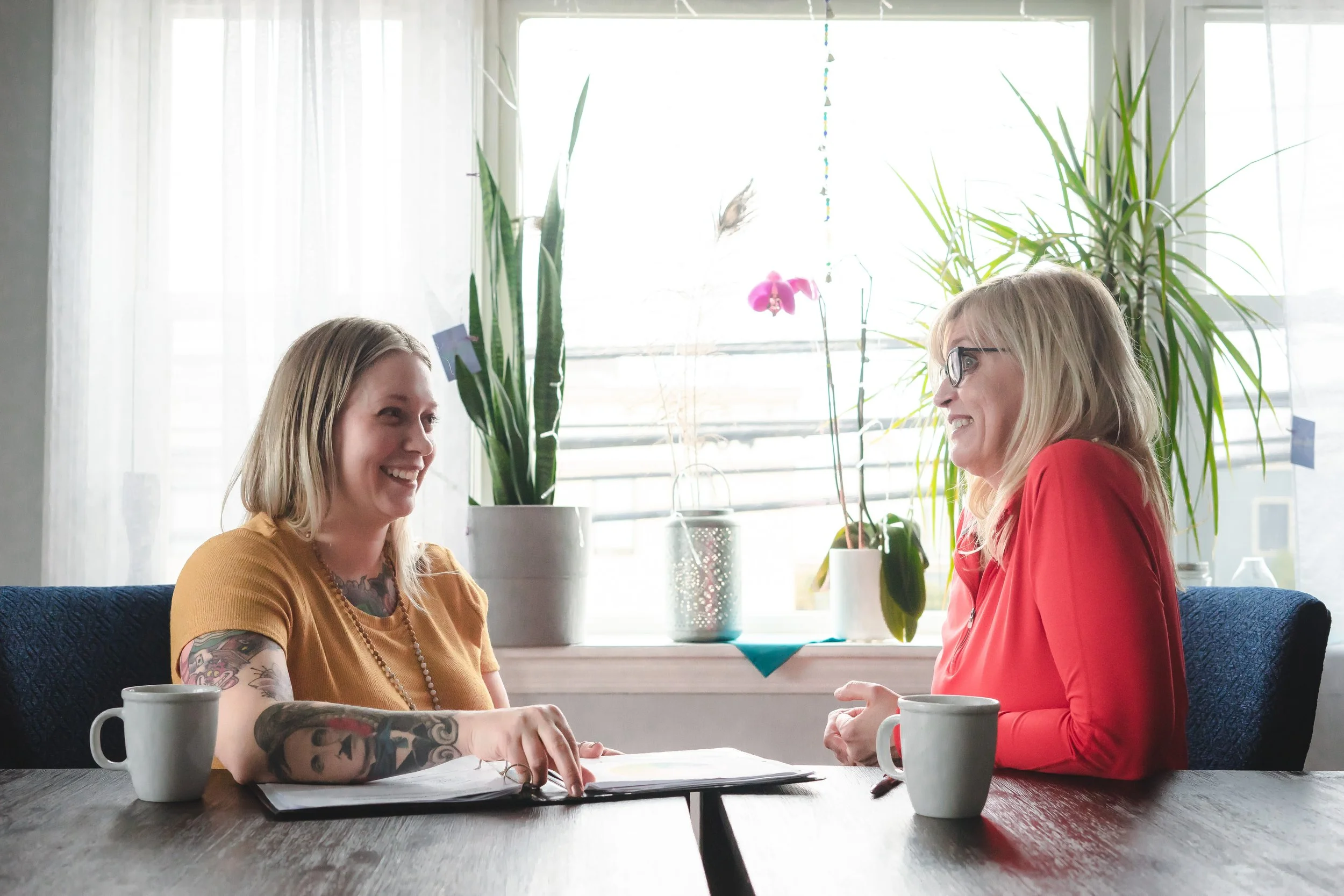 Two women sitting at a table having a conversation in a bright room with large windows, plants, and coffee mugs.