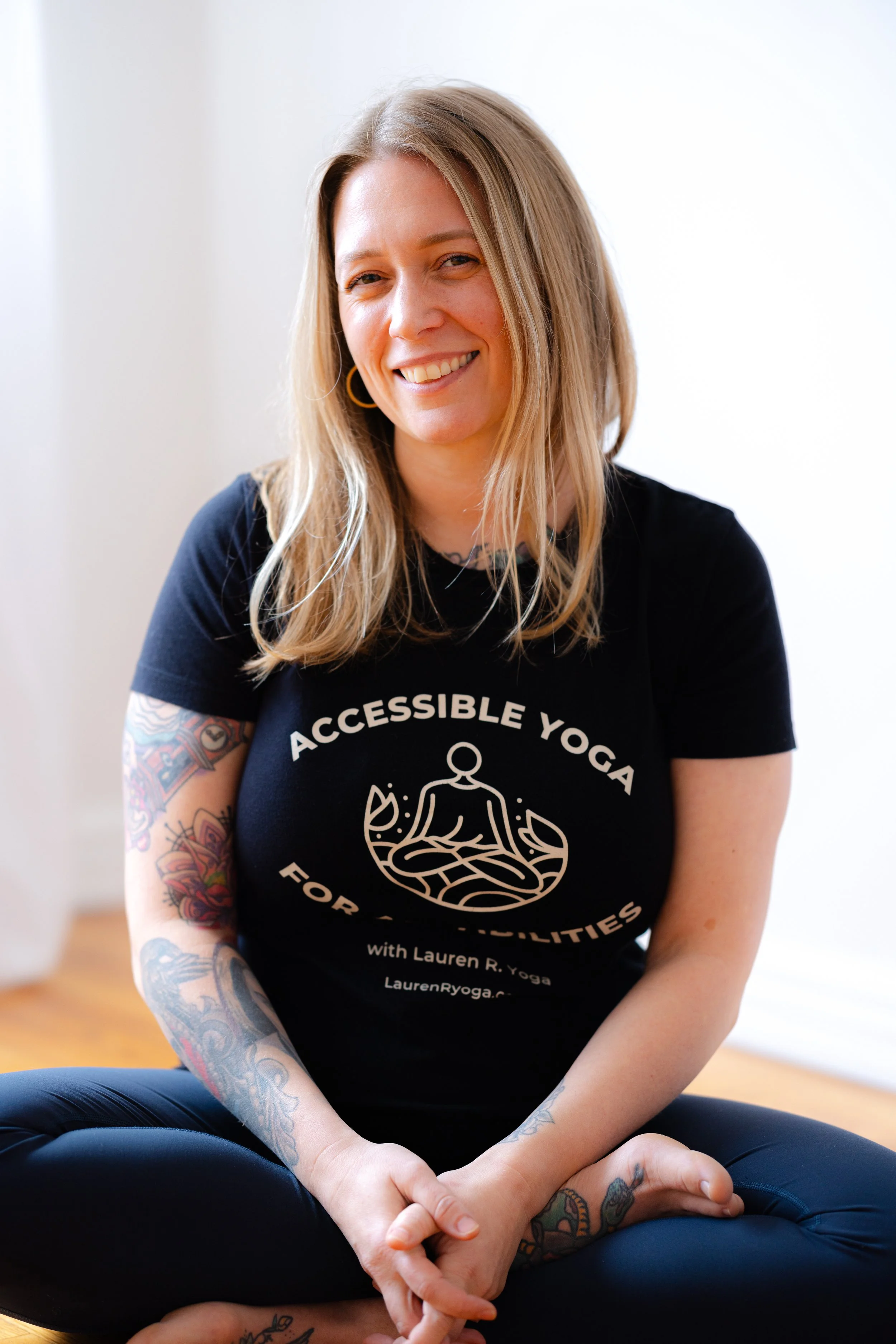 Lauren Reek private yoga teacher Long Island sitting cross legged smiling wearing a black t-shirt with text about accessible yoga for all abilities