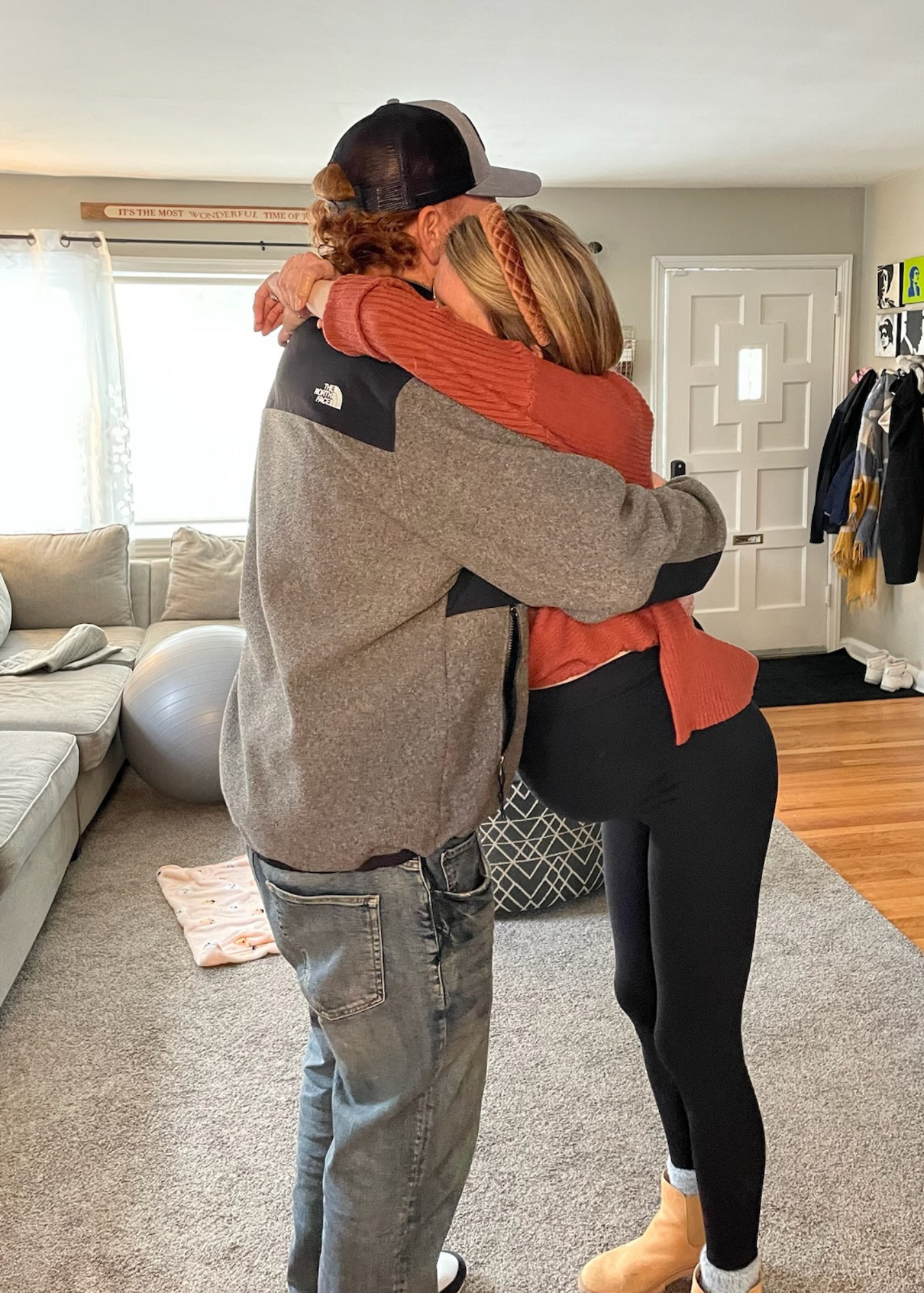 A man and a woman are hugging each other warmly inside a living room with a beige couch, a window, a door, and wall decorations visible in the background.
