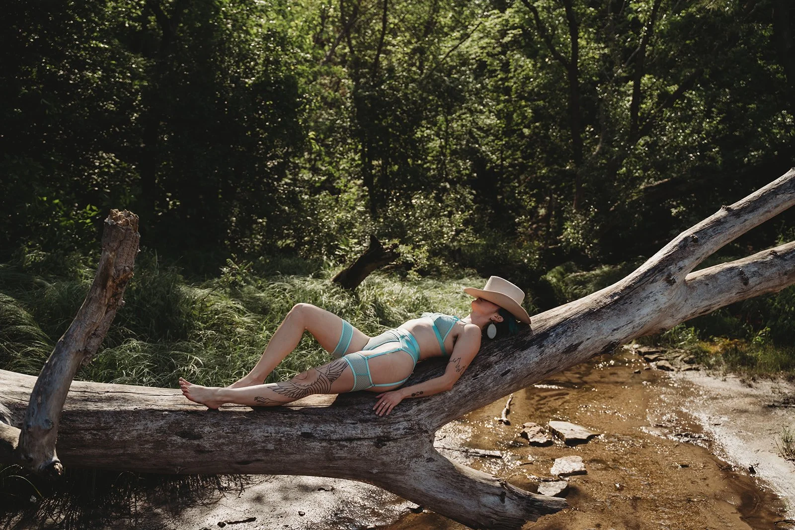 nonbinary queer person with tattoos wearing blue lingerie and laying on a fallen tree with a cowboy hat over their face